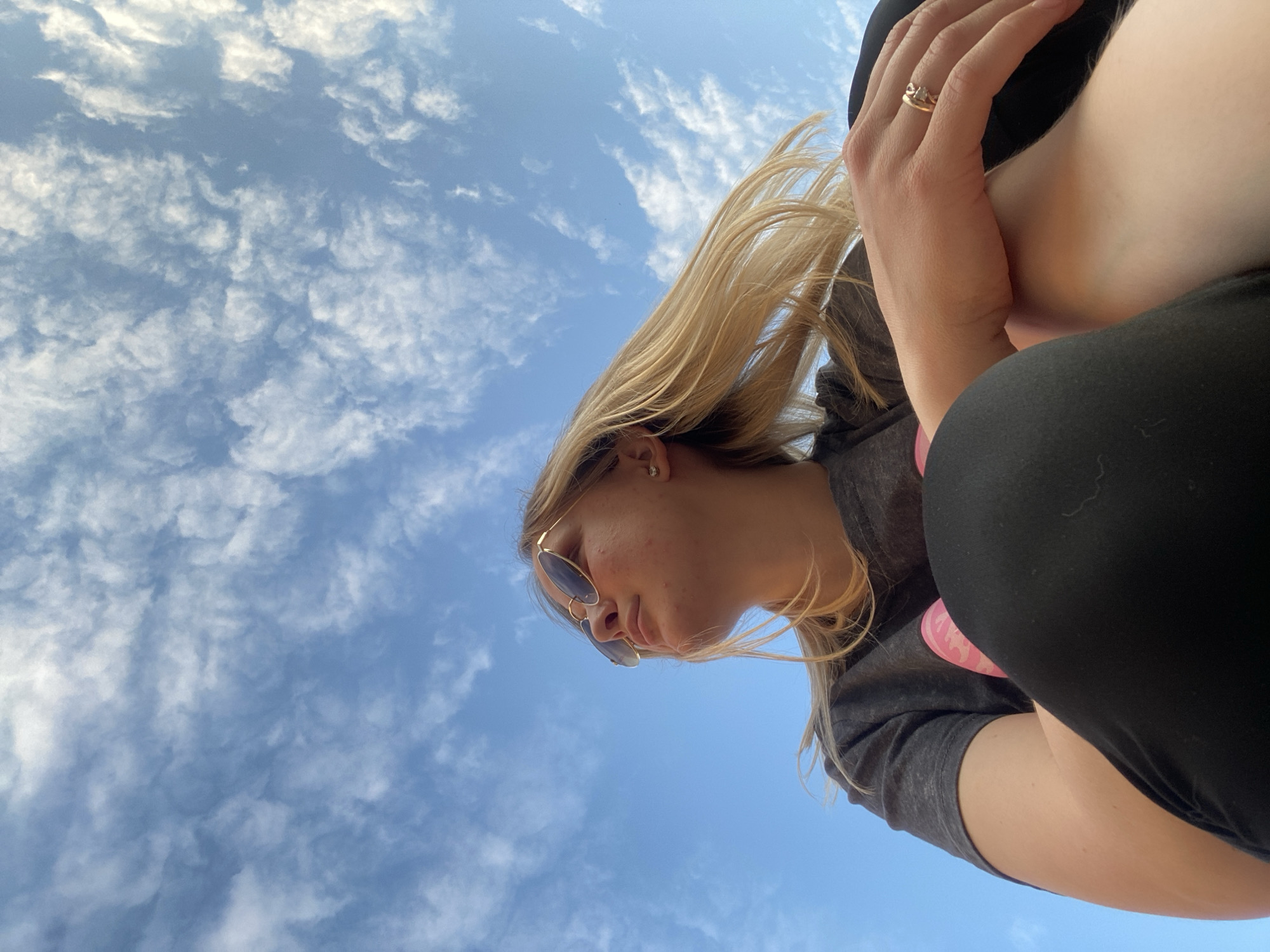A low-angle shot captures an individual against a blue sky with scattered white clouds. The person, identifiable as female with blonde hair, fair skin, and a few small blemishes on her cheek, is positioned with her head tilted upwards and slightly to the left, looking into the distance. She wears sunglasses with gold frames. A small, clear stud earring is visible in her left ear. Her attire consists of a dark grey t-shirt and black bottoms, likely pants or leggings. Her right hand, adorned with a gold ring featuring a clear stone on the ring finger, rests on her bent knee. The overall composition is from a worm's-eye perspective, emphasizing the vastness of the sky above the subject. The location is Unknown, Unknown.