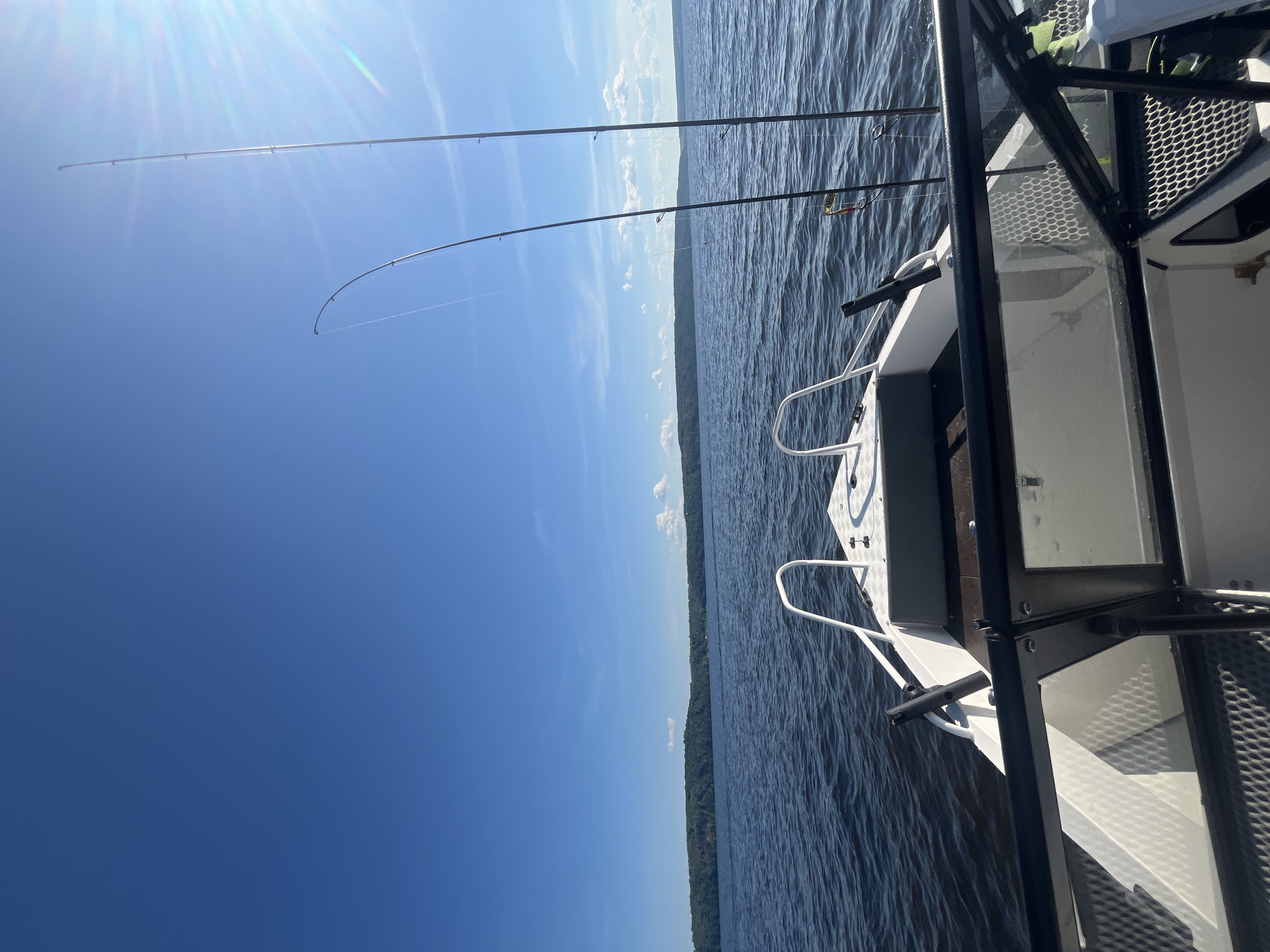 The image shows a view from the perspective of someone inside a boat on a body of water. The boat has white railings and a windshield. Two fishing rods are prominently placed on the right side of the boat, with fishing line visible. The water is a deep blue and appears to be slightly choppy. In the distance, a tree-covered shoreline is visible under a bright blue sky with scattered white clouds and the sun shining from the upper right, creating lens flare. The scene is consistent with a location on a lake or a large river in Siukterka, Russia, likely during daylight hours.