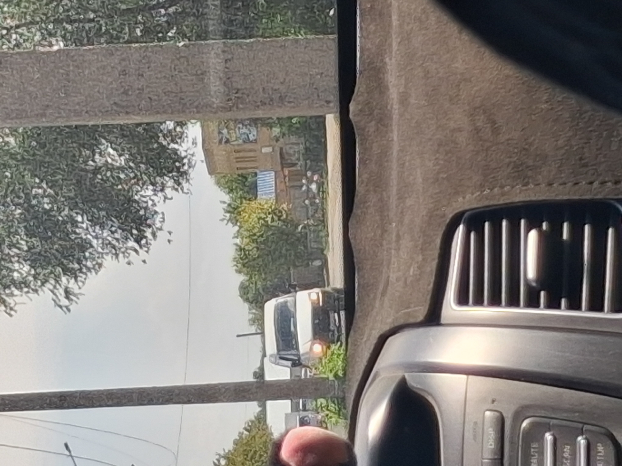 A white van is parked on a dirt road with a building and trees in the background. The photo was taken from inside a car, with the dashboard visible in the foreground.