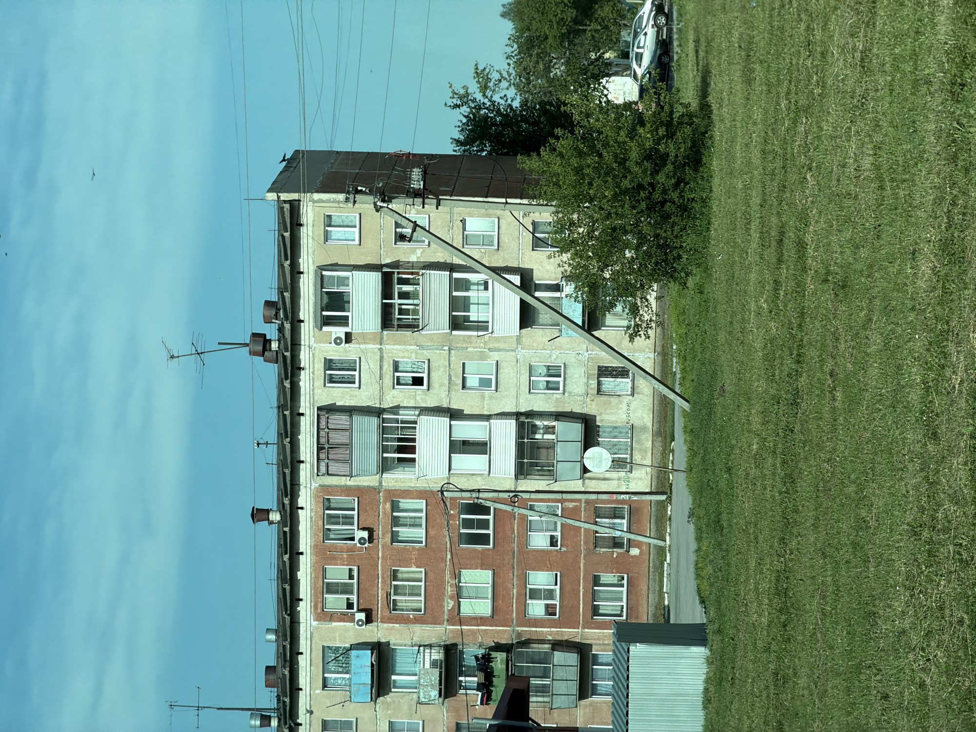 The image depicts a multi-story apartment building in Yevsino, Russia, under a bright, clear blue sky with a few wispy clouds. The building, likely from the Soviet era, features a faded, textured facade with alternating sections of light beige and reddish-brown. Numerous windows, many with white frames, punctuate the building's exterior, some with balconies enclosed with glass or featuring railings. Air conditioning units are visible mounted below some windows.
The building's roof is flat and appears somewhat weathered, with several metal chimneys and ventilation pipes protruding from it, alongside antennas. Utility wires crisscross the sky above the building. A large, concrete utility pole runs diagonally across the facade, supporting a network of electrical cables and what appears to be a transformer box. A circular sign is affixed to this pole.
In the foreground, a well-maintained lawn of green grass stretches out, appearing slightly uneven with some areas of dryer, lighter-colored grass. To the left of the apartment building, a small, metallic shed-like structure with ribbed siding is visible. Behind the building and to the right, a cluster of lush green trees provides a contrast to the urban architecture. A light-colored car and a darker car are parked on a paved area behind the trees. The overall scene suggests a typical residential area in a Russian town.