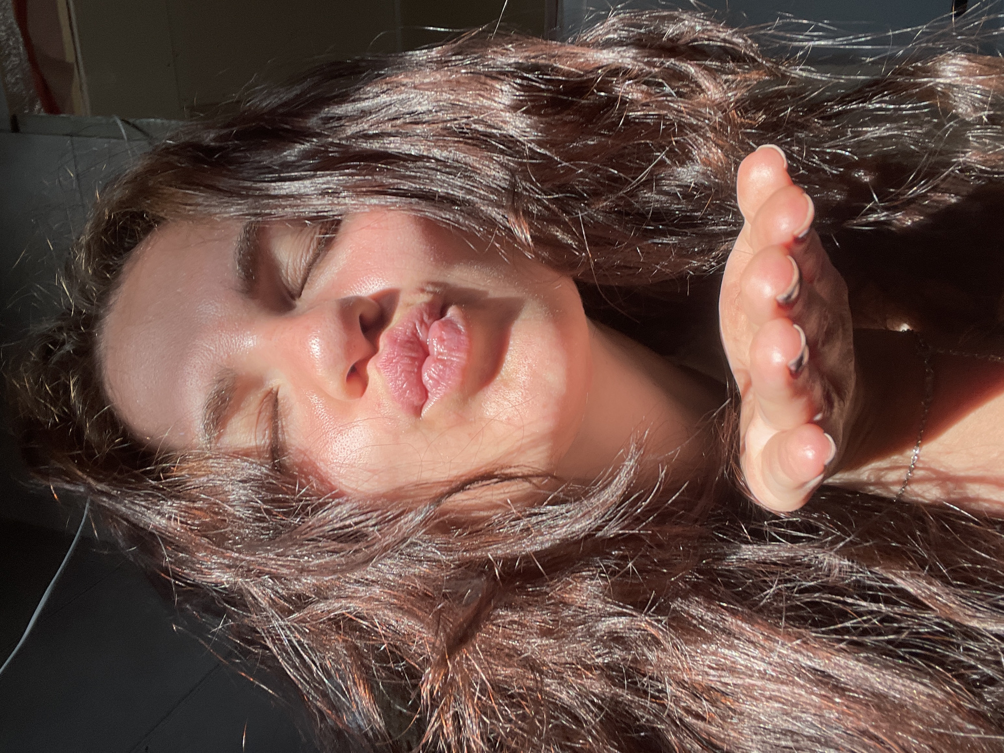 A close-up selfie shows a young woman with long, dark brown, wavy hair. She is wearing a dark shirt and a thin silver chain necklace. Her eyes are closed, and her lips are puckered as if blowing a kiss. Her right hand is extended forward, palm up, with her fingers slightly curled. Sunlight illuminates her face, highlighting her skin and the texture of her hair. The background is dark and appears to be indoors, possibly a room with industrial elements suggested by pipes or conduit visible at the top right. The image is well-lit, with strong contrast between light and shadow. There is no indication of the specific location within Мосметаллоконструкция Онт, Russia.