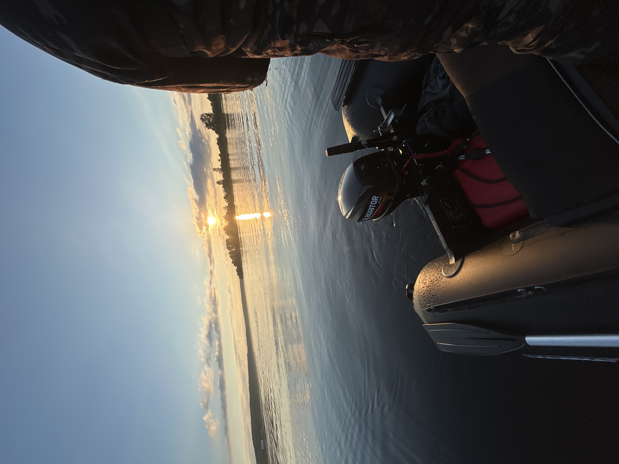 A person wearing camouflage clothing is sitting in a black inflatable boat on a calm body of water. The sun is setting on the horizon, casting a warm glow across the sky and water. Trees and a shoreline are visible in the distance. A small outboard motor is attached to the back of the boat, and a red gas can is secured inside. The water is dark and reflective, with small ripples on the surface. An oar is propped up against the side of the boat. The location is Ol'khovskoye, Russia.