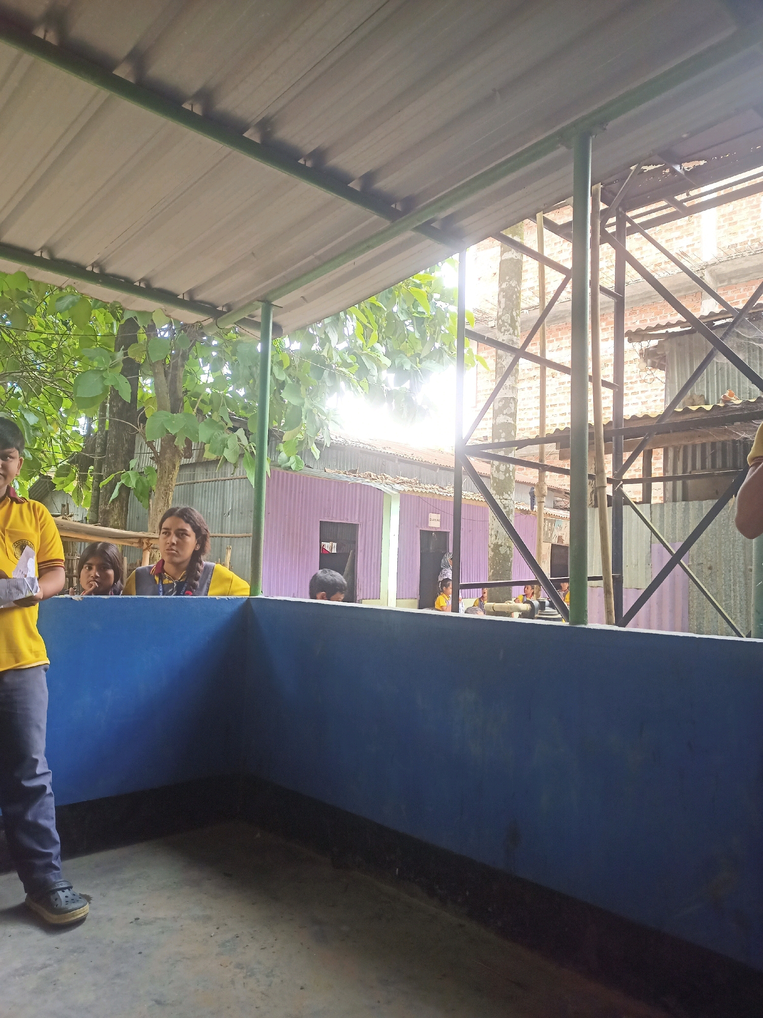 This image depicts an outdoor setting, likely at a school in Hatsinigmari, India, with a group of children and young people visible. The foreground features a blue-painted wall or barrier. To the left, a young boy wearing a yellow shirt and dark pants stands holding a white object. Behind him, two younger girls are looking towards the camera. Further back, a teenage girl with braided hair, wearing a yellow and gray shirt, is also looking forward. In the background, there are various structures including corrugated metal roofs, brick walls, and a metal frame. A purple building with a dark doorway is also visible. Sunlight streams in from the right, creating bright highlights and casting shadows. The overall impression is a candid moment captured during an outdoor activity or gathering.