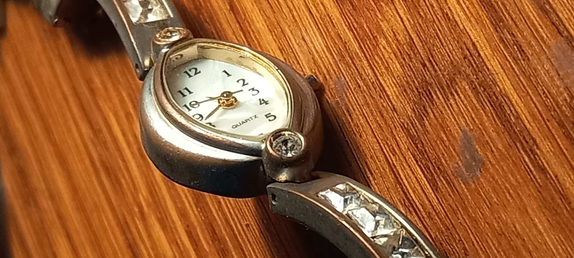 A close-up, eye-level shot shows a ladies' silver-toned quartz wristwatch with a white mother-of-pearl dial resting on a wooden surface. The watch features a oval-shaped case with a beveled edge. The dial has black Arabic numerals and gold-toned hour and minute hands. The word "QUARTZ" is printed on the lower half of the dial. The watchband is silver-toned and has decorative square-shaped clear stones set into it. Two small clear stones are visible on the lugs of the watch where the band connects to the case. The wooden surface appears to be a table or countertop with a visible wood grain pattern, oriented diagonally in the frame. The lighting creates a slight shadow behind the watch. The image was taken in Kryvyi Rih, Ukraine.