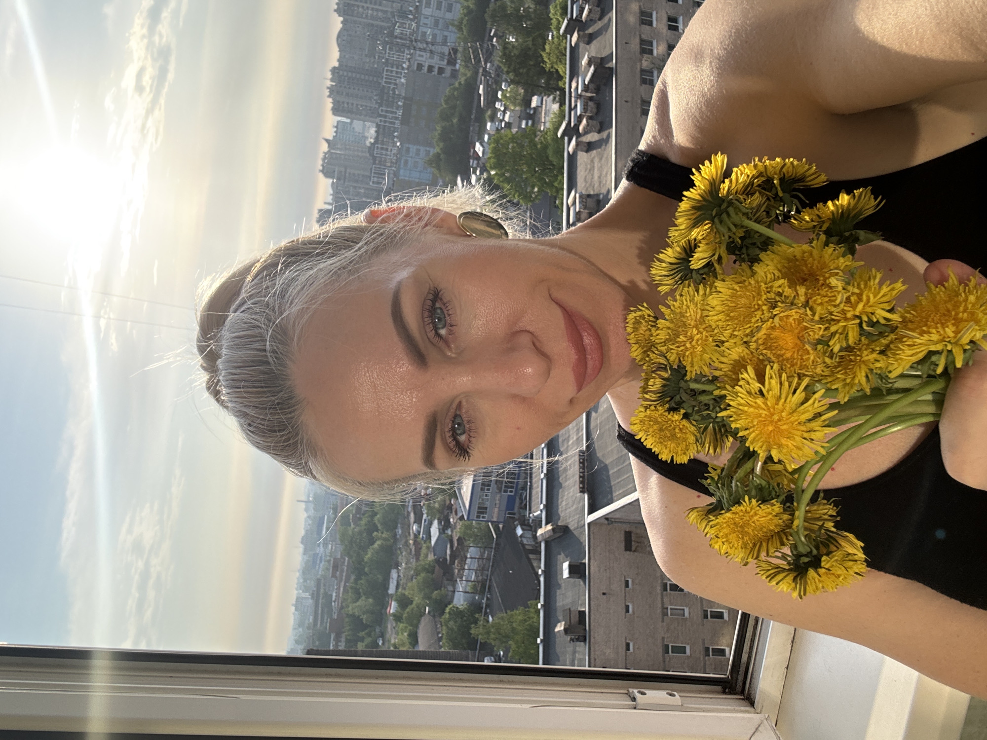 A woman with blond hair pulled back in a bun stands inside by a window, holding a bouquet of dandelions. The background shows a cityscape with buildings, trees, and a hazy sky. The location is in the city of Unknown, Unknown.