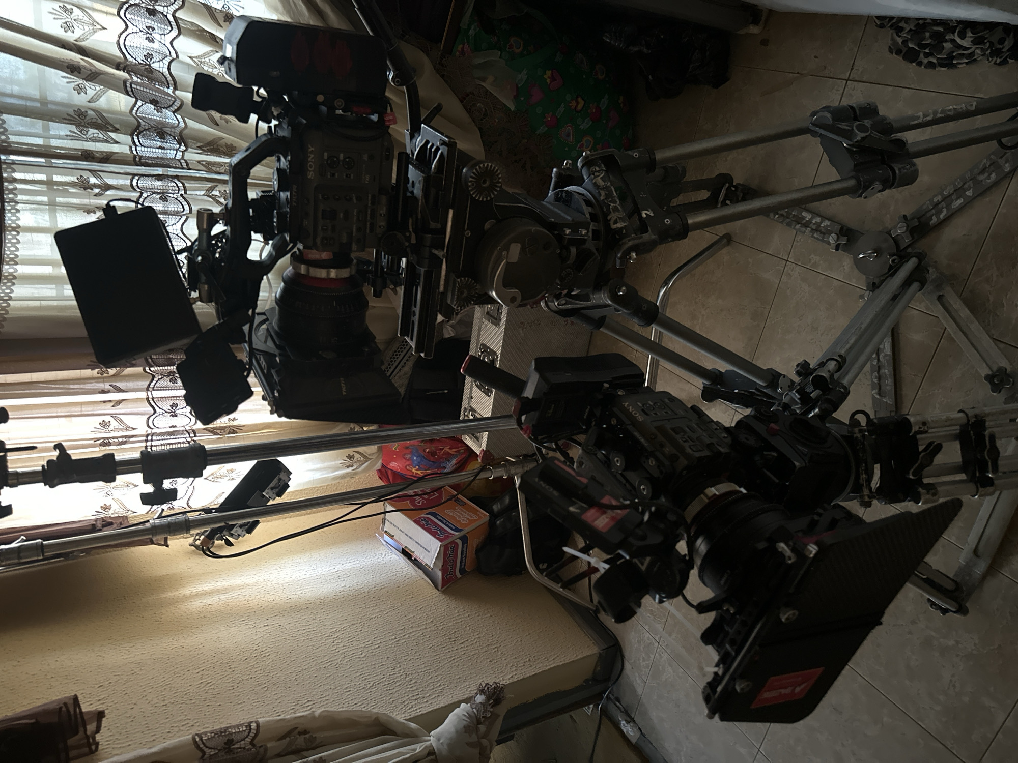 The image, rotated counter-clockwise, presents two professional video camera setups in an indoor environment, likely a production set or studio. In the foreground, a sophisticated Sony cinema camera is mounted on a sturdy tripod, featuring a large lens, a matte box, and an attached external monitor. Various accessories, including what appears to be a Tilta rig, are integrated into its setup.
Further back and to the right, a second, similarly equipped Sony cinema camera is positioned on a camera slider or dolly system, which itself is supported by another tripod. Both cameras are rigged for filming, indicating an active or preparatory stage of a video production.
The background features translucent curtains with decorative patterns, through which natural light enters, suggesting it is daytime. The floor is covered with light-colored square tiles. Several items are scattered around the camera equipment on the floor, including a distinctive cardboard box of "Indomie Instant Noodles," specifically "Fried Noodles Mi Goreng." Other bags and fabric items are also visible.
Visible text includes "SONY" on the camera bodies, "TILTA" on some of the accessories, and "RED" on a small tag attached to one of the camera rigs. The "Indomie" box clearly shows its brand name and product description. The scene conveys a professional film or video production setting in Lagos, Nigeria, given the product placement. No individuals are present in the image.