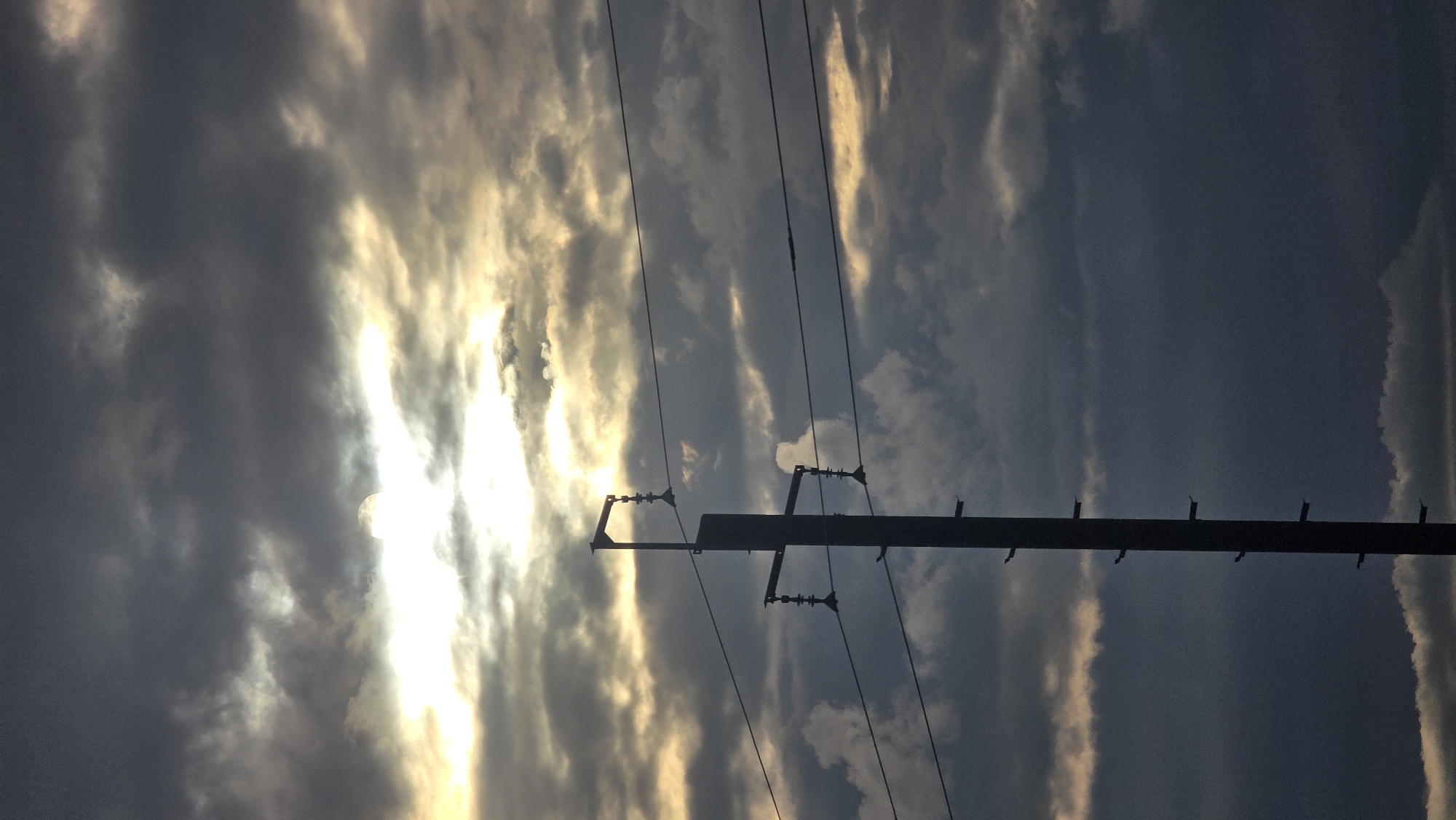 The image displays a tall, dark utility pole, likely in Leninogorsk, Russia, standing in stark silhouette against a heavily clouded sky. The pole, positioned on the left side of the frame and extending from the bottom upwards, features multiple cross arms or brackets from which bell-shaped insulators are visible, supporting several taut power lines that stretch horizontally across the middle ground. Smaller, ladder-like attachments are present on the right side of the pole. The sky is dominated by dense, dark grey and blue-grey clouds, yet a brilliant, golden-white light source, consistent with a partially obscured sun, radiates intensely from the upper-left to the central part of the frame, illuminating the edges of some clouds with a golden hue and creating significant contrast. No individuals, animals, or explicit actions are depicted within the scene.