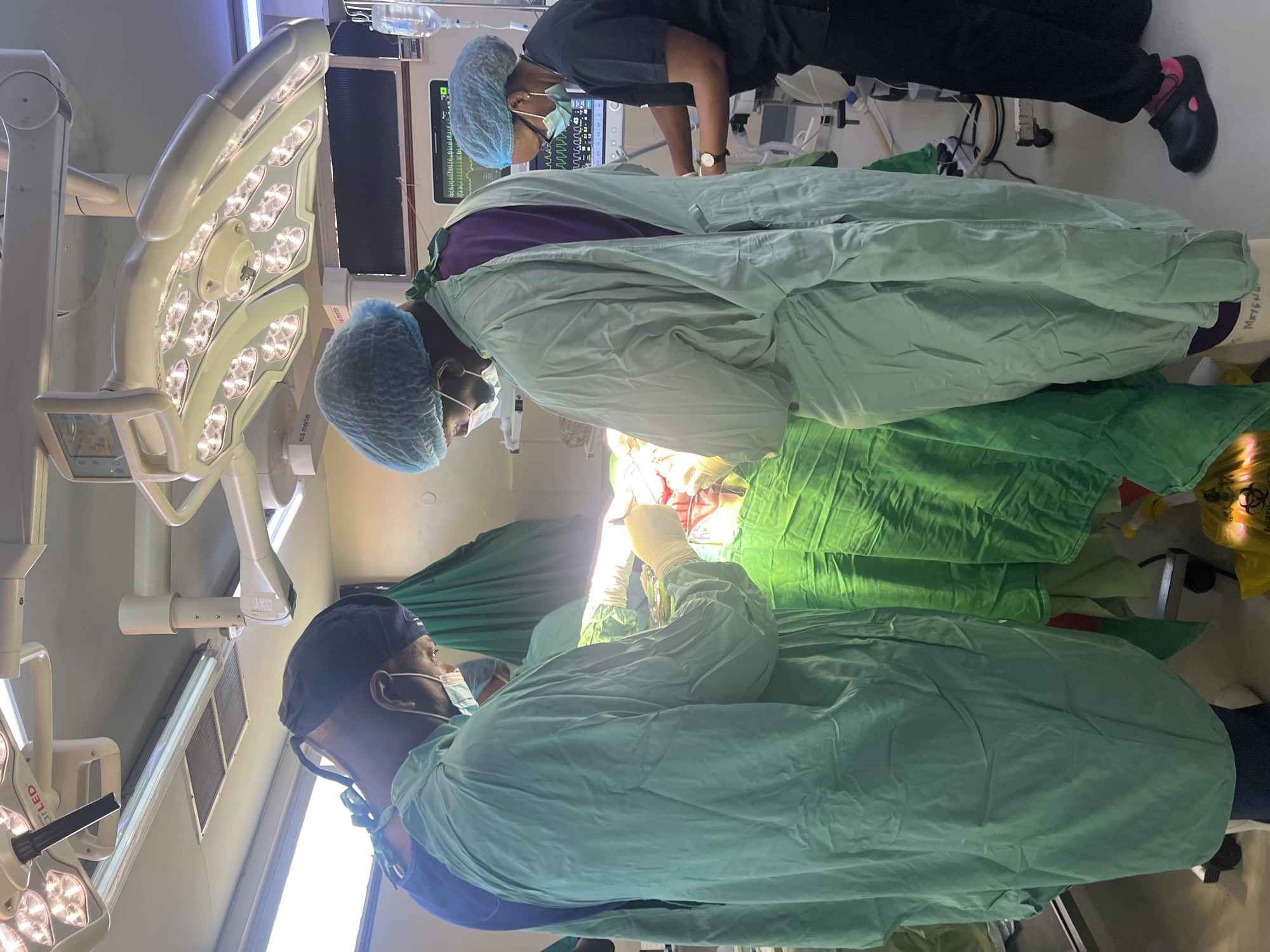 The image depicts an operating room scene, likely in Nairobi, Kenya. Three individuals are visible, all wearing surgical scrubs, caps, and masks, suggesting they are medical professionals. Two are actively engaged in a surgical procedure on a patient who is draped with green surgical sheets. The third individual appears to be monitoring equipment, including a vital signs monitor displaying various waveforms and numerical values. A surgical light fixture is prominently visible above the surgical site. Additional medical equipment, including an IV bag and various cables, can be seen in the background. The environment is a sterile operating room setting with white walls and ceiling.