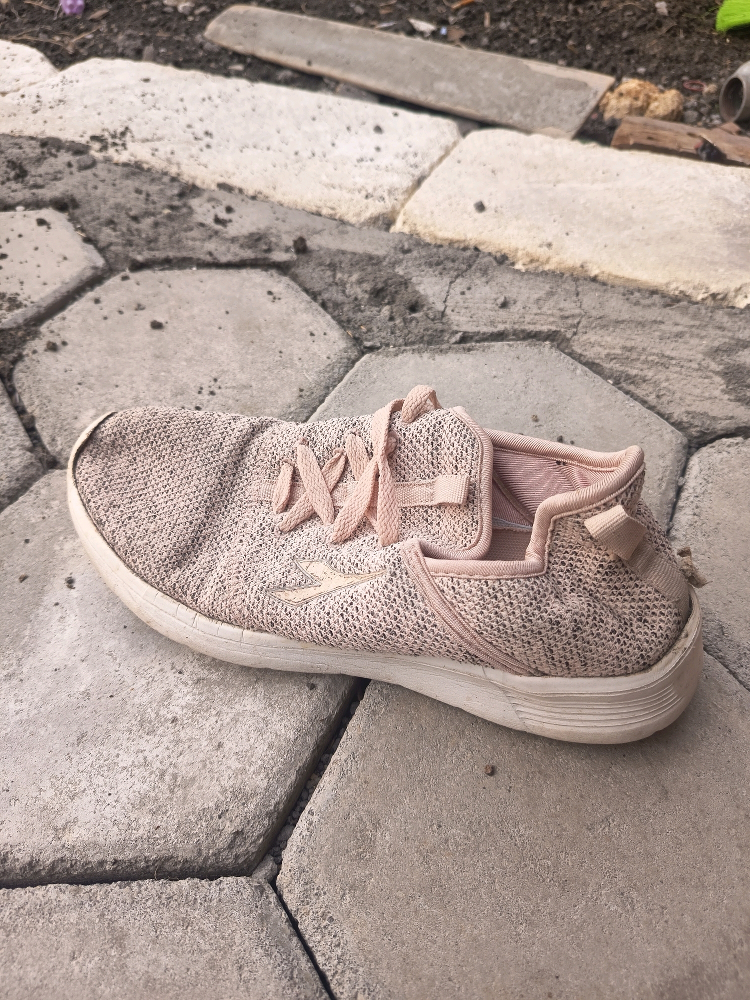 The image shows a single, pink, woven athletic shoe lying on a textured, grey, hexagonal pavement. The shoe is positioned on its side, with the sole facing upwards. The pavement is outdoors, with dirt and small debris visible in the background. The scene appears to be an unkempt area, possibly a pathway or yard. There are no people or any other discernible objects in the immediate vicinity of the shoe. The lighting suggests it is daytime. The specific location in Sidorejo, Indonesia cannot be determined from the visual information provided.