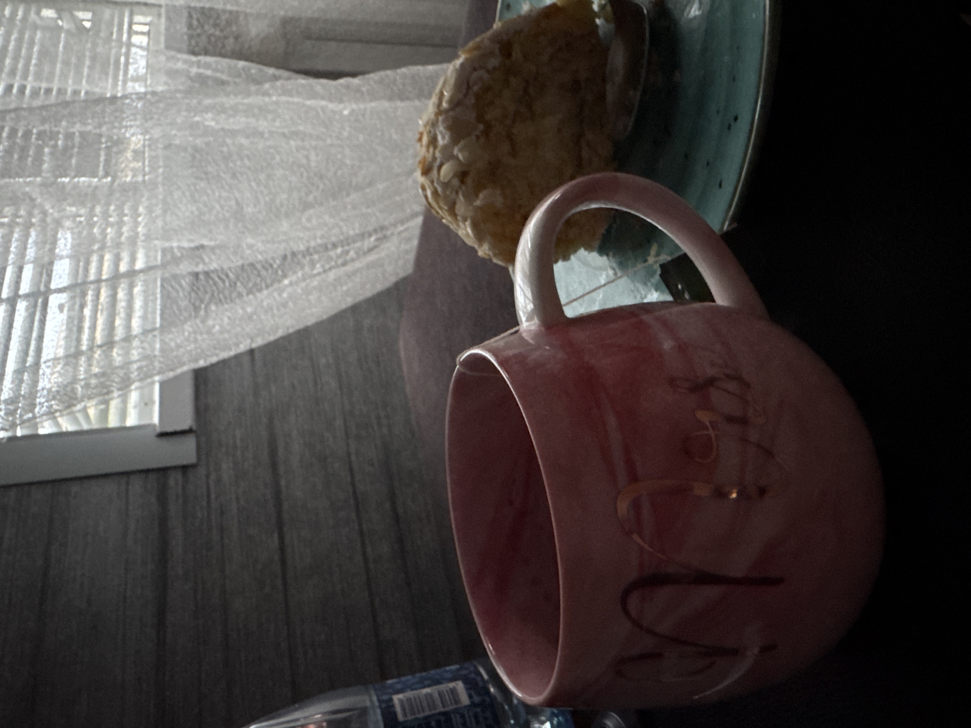 A pink mug with "Mrs" inscribed in gold script sits on a dark surface. Next to it, on a teal-colored plate, is a pastry with a textured, crumbly topping. A bottle of water is partially visible to the left of the mug. In the background, a window with horizontal blinds is covered by sheer white curtains. The overall scene appears to be indoors, possibly in a home or cafe.