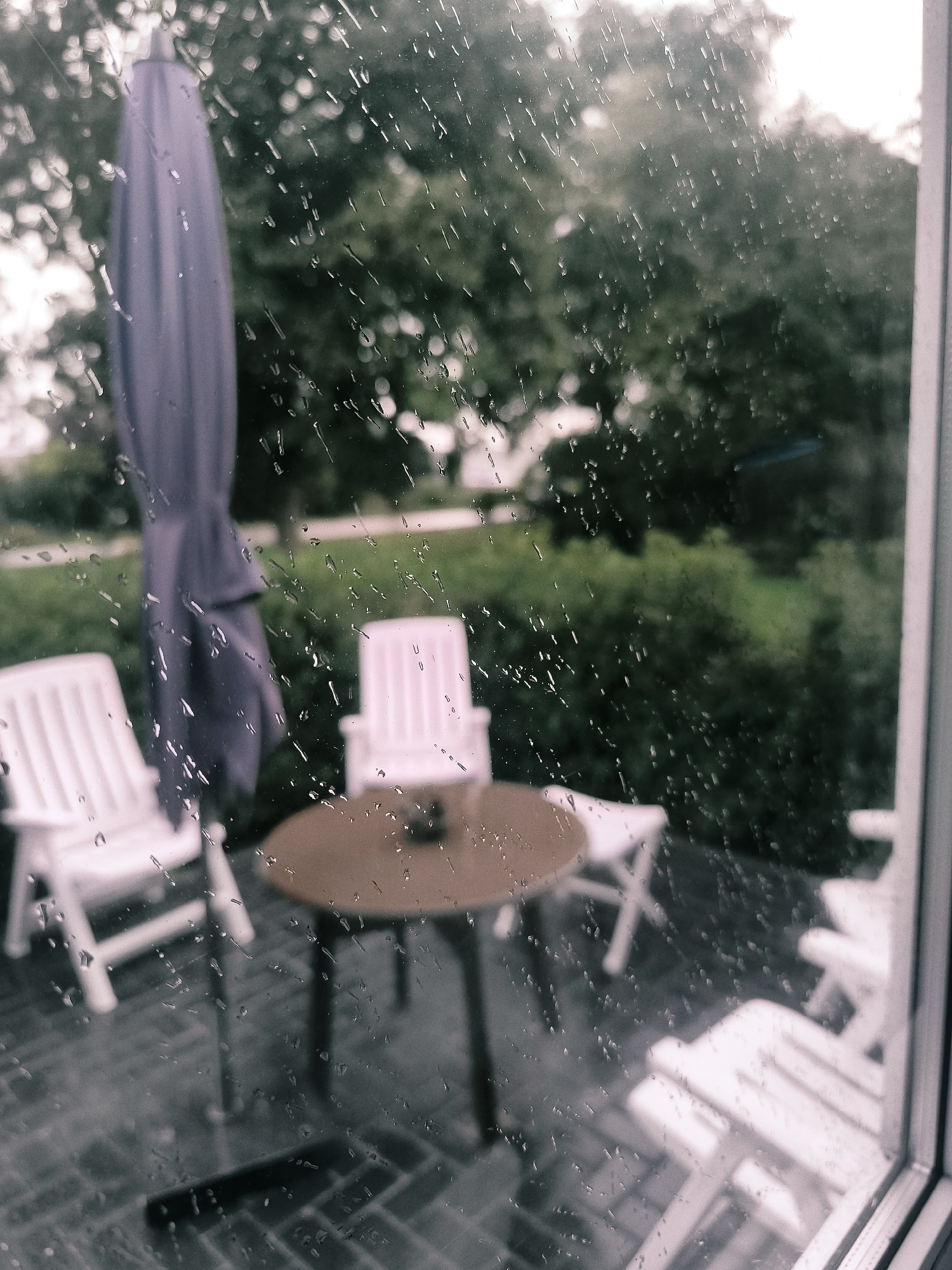 The image, taken through a rain-streaked window, depicts an outdoor patio area. Visible are two white plastic lounge chairs, a round brown table with an object on it, and a closed purple umbrella. In the background, lush green trees and bushes form a natural backdrop. The ground appears to be paved with dark grey bricks or tiles. The overall atmosphere suggests a tranquil, possibly suburban or residential setting in Almere, Netherlands, experiencing inclement weather. There are no people visible in the scene.