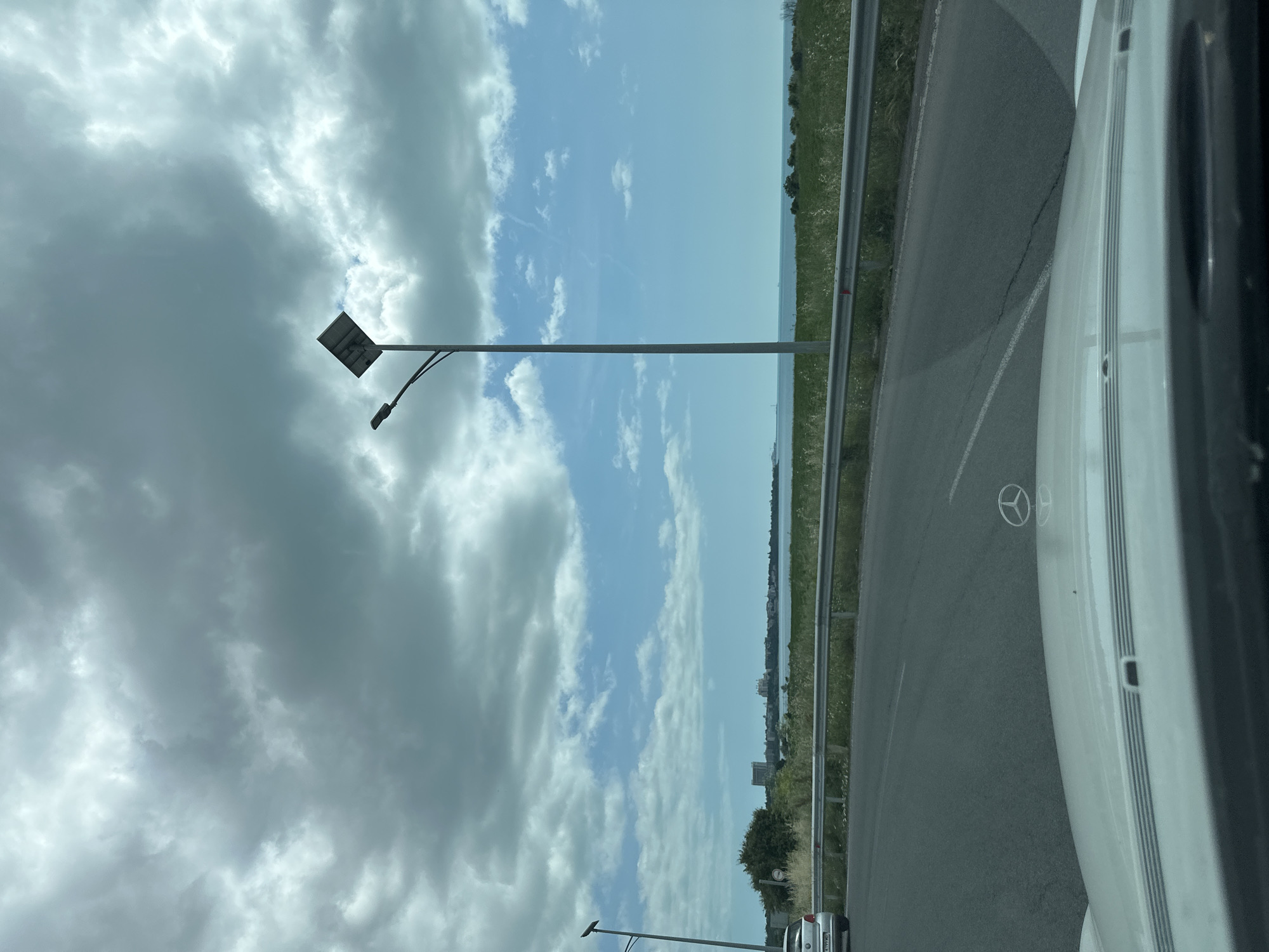 A wide-angle, eye-level shot captures a view from a vehicle in Pomorie, Bulgaria, looking towards the Black Sea under a cloudy sky. In the foreground, the hood of a white car with a Mercedes-Benz emblem is visible. A paved road curves to the left, bordered by a metal guardrail and a grassy area with sparse vegetation. A solar-powered street lamp stands tall on the right side of the road, its panel angled towards the sky. In the middle ground, a line of trees and buildings forms a distant coastline. The horizon is defined by the calm sea and a sliver of land. The sky is filled with dramatic, gray clouds interspersed with patches of bright blue sky and wispy white clouds. To the left, another street lamp is partially visible, and a silver car is parked on the side of the road.
