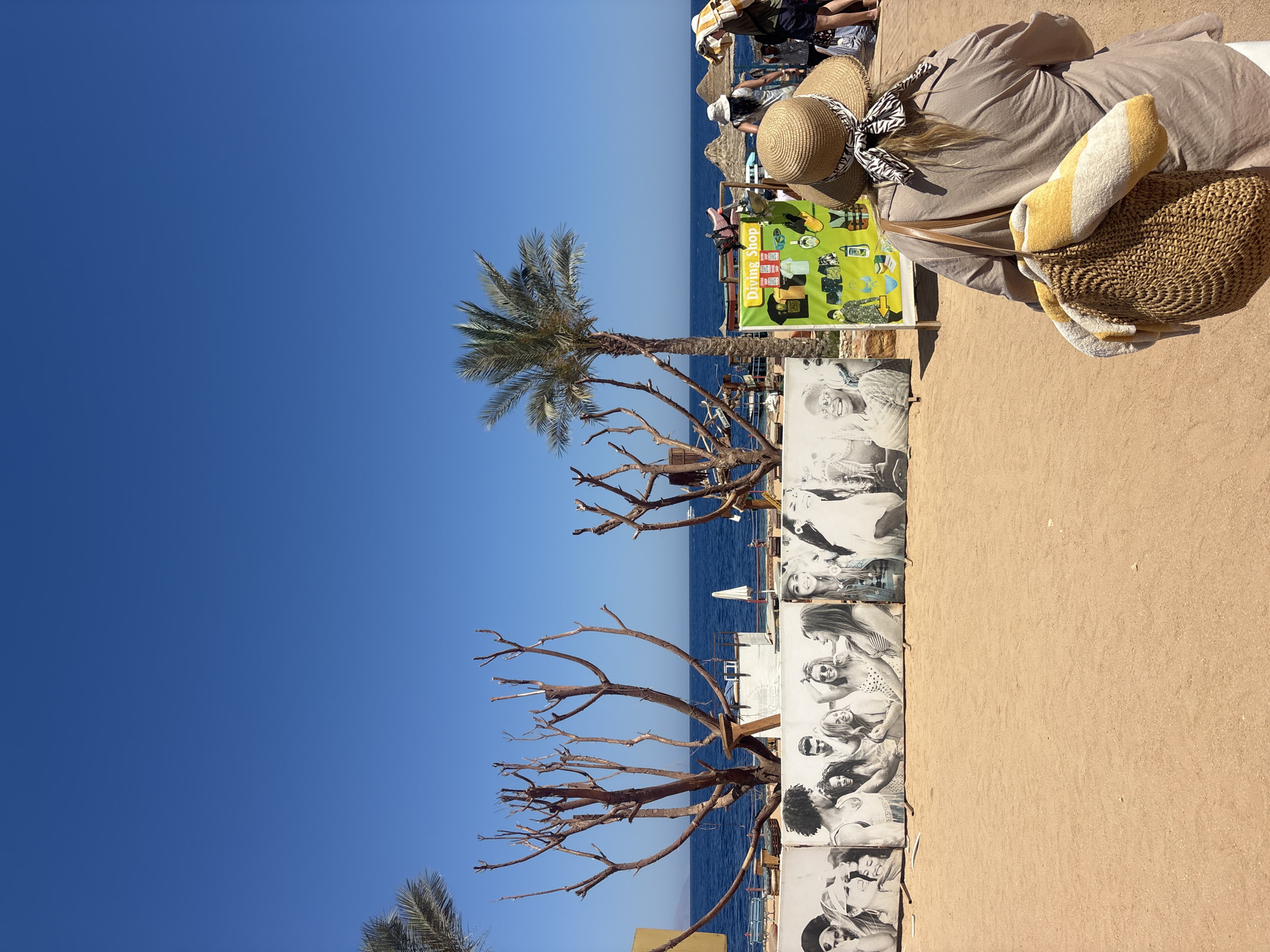 The image captures a scene on a beach. A person is lying down wearing a sun hat, a straw bag with a striped towel, and a cover-up. To the left, there is a diving shop sign. Further left, a palm tree is visible, along with a bare tree. In the background is the ocean, a sandy beach, and a clear blue sky. The location appears to be a beach in an unidentified city.
