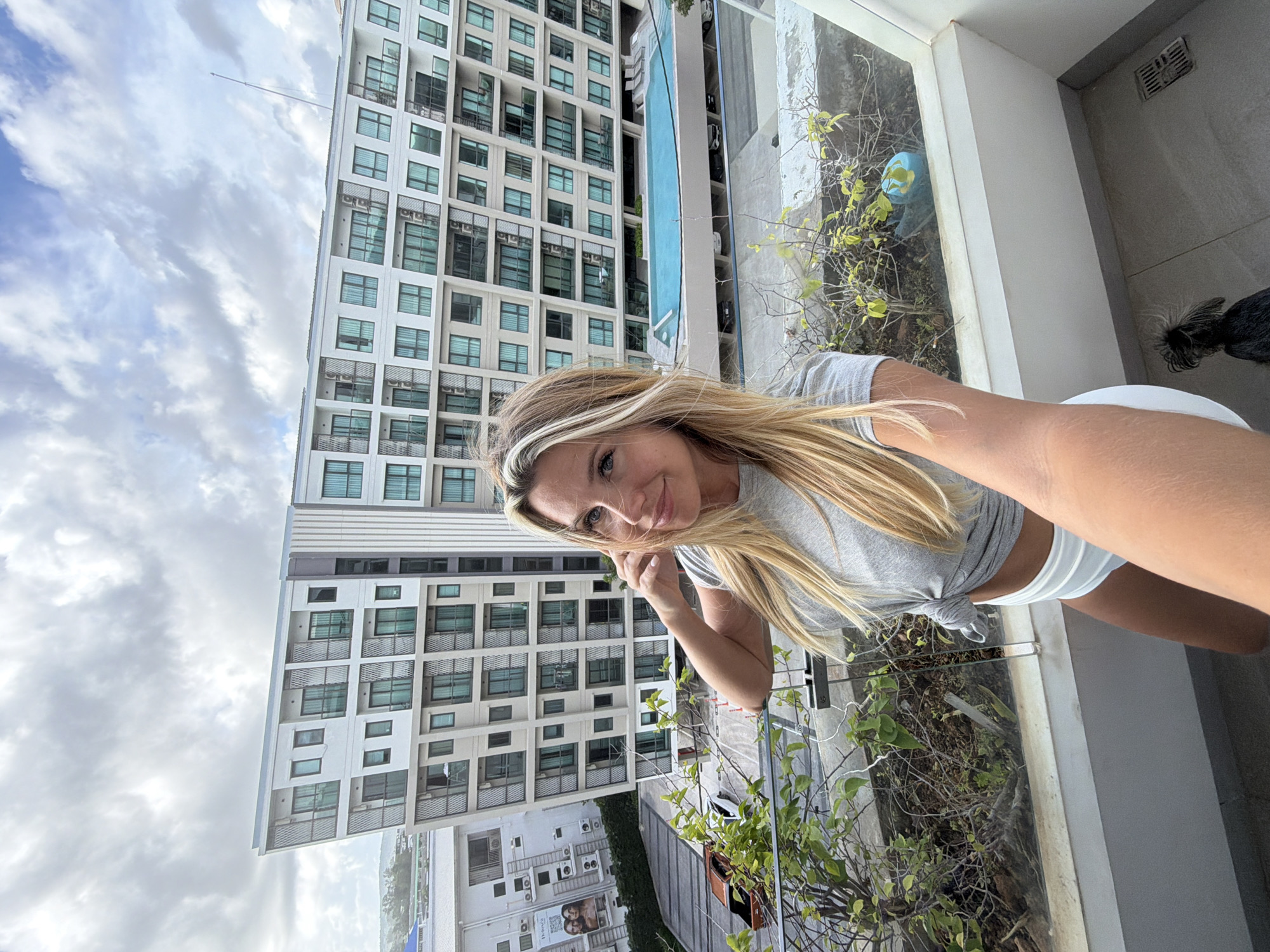 A blonde woman with blue eyes is taking a selfie on a balcony overlooking a residential building in Choeng Thale, Thailand. The woman is wearing a grey t-shirt and white shorts. She is standing on a balcony with a glass railing and plants. In the background, there is a large modern building with many windows and balconies, and a swimming pool. The sky is cloudy. A small black dog is visible on the floor of the balcony.