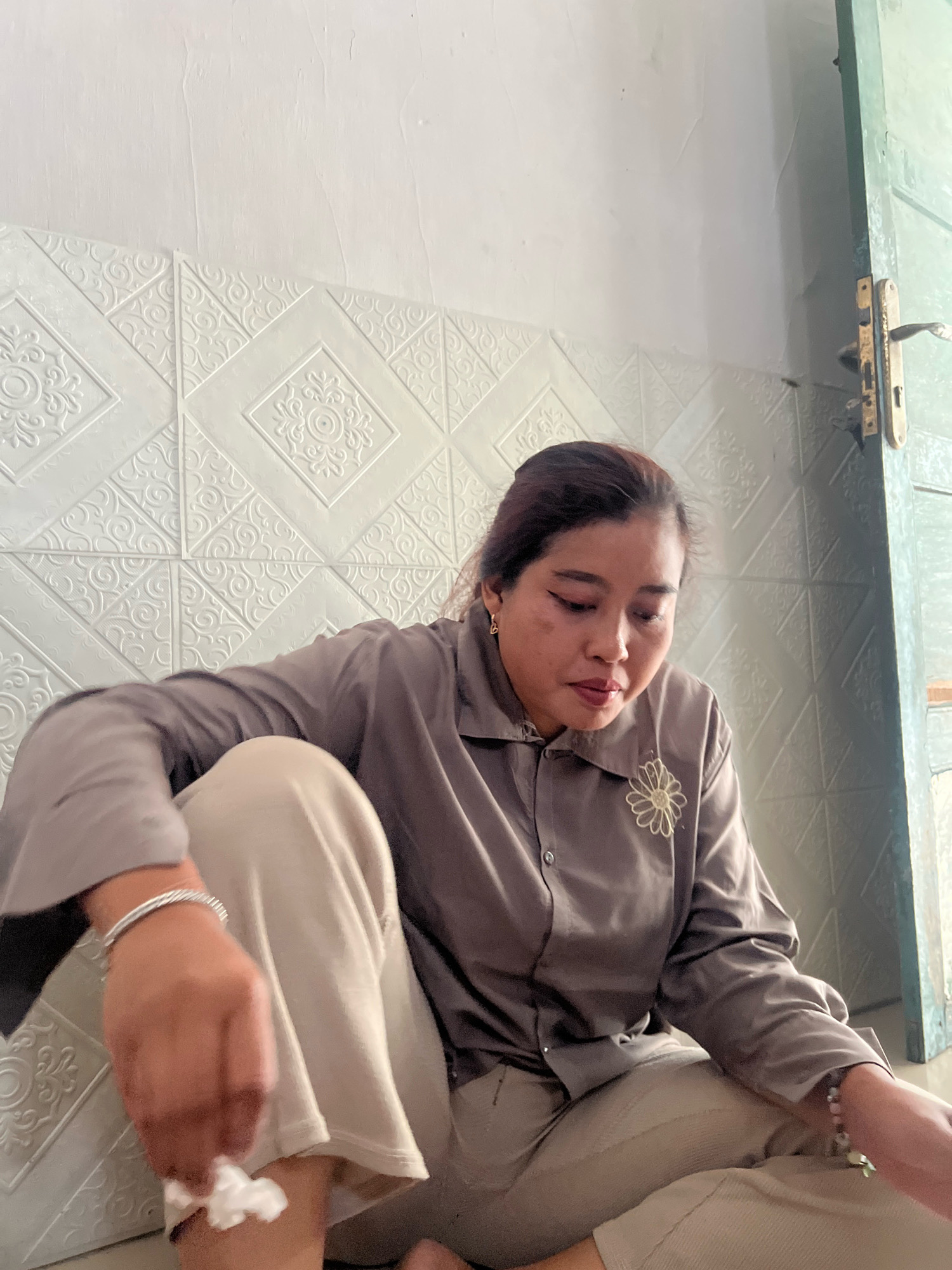 A woman, possibly in her 30s or 40s, is seated on the floor of a room with textured white walls. She is wearing a loose-fitting, long-sleeved brown shirt with a small embroidered flower on the left breast and light-colored pants. Her hair is pulled back, and she has visible makeup. A silver bracelet is on her left wrist, and she is holding a crumpled piece of white material in her right hand. To the right of the woman is a partially visible green door with a brass handle. The background wall features a repeating geometric pattern in relief. The scene appears to be indoors, likely in a residential setting in Kecamatan Pati, Indonesia, given the context.