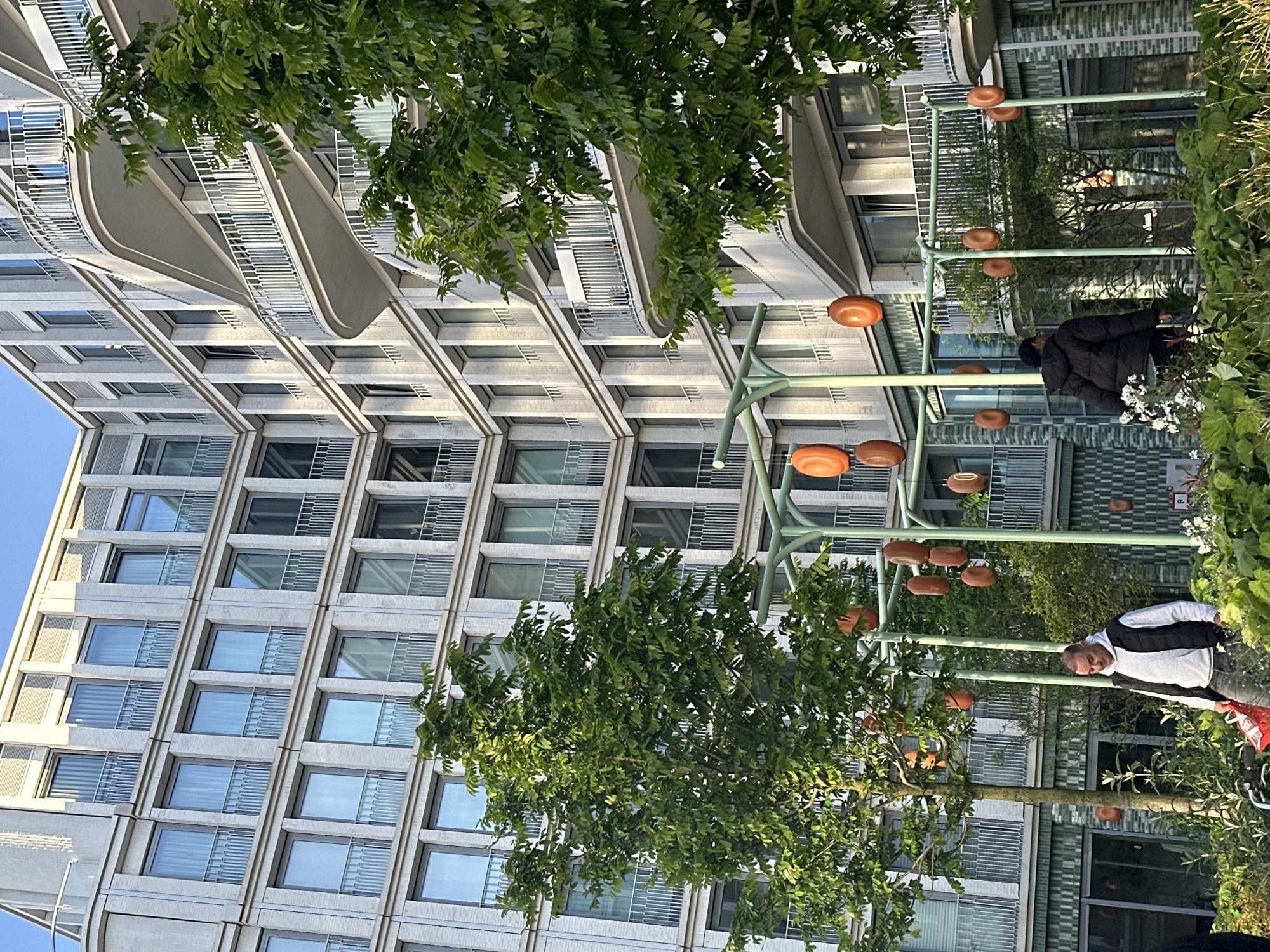 The image captures an outdoor scene in Amsterdam, Netherlands, featuring modern architecture and urban greenery. A tall, contemporary building with a grid-like facade and curving architectural elements dominates the left side of the frame. Windows, some with metal screens, are regularly spaced across the building's surface. A lush tree with dense green foliage obscures parts of the building.
In the foreground, a series of light green metal structures with evenly spaced, disk-shaped orange lights extend across the scene. These structures create a canopy-like effect. Two individuals are visible. One person, wearing a black jacket, is seen near white flowers and vegetation. Another individual, wearing a black and white jacket, is walking with a red bag or object.
The environment includes a blend of natural and urban elements. Vegetation, including trees and plants, adds a layer of greenery to the scene. The architecture, lighting, and human activity create a snapshot of urban life in Amsterdam.