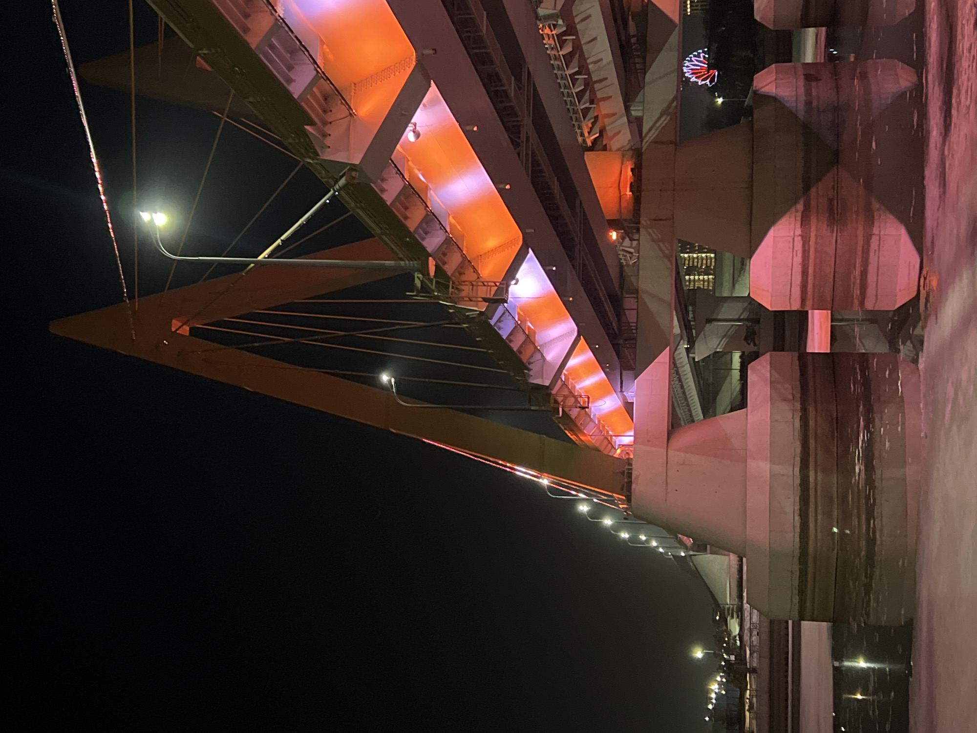 The image captures a nighttime view of the Millennium Bridge in Kazan, Russia, spanning the partially frozen Kazanka River. The cable-stayed bridge features prominent orange A-shaped pylons and a structural deck illuminated primarily by integrated orange and purple lighting. Multiple streetlights are mounted along the bridge deck, emitting bright white light. The water below the bridge contains ice floes and reflects the vivid colors of the bridge's illumination. Several large concrete piers supporting the bridge superstructure are visible emerging from the icy water. In the distant background, discernible city lights mark the urban landscape, and a brightly illuminated Ferris wheel with red and white elements is partially visible on the far right. No individuals, vehicles, or specific human activities are observable within the frame.