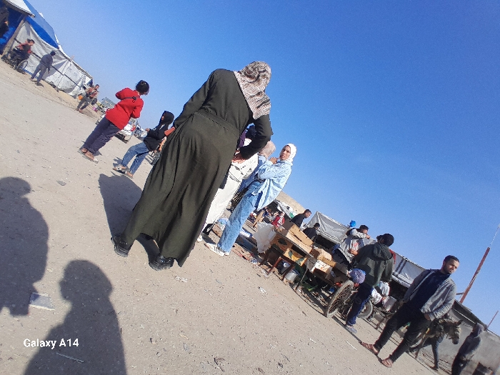 The image captures an outdoor, daytime scene under a clear blue sky in Khan Yunis, Palestinian Territory. The ground is unpaved, appearing as dirt or gravel. In the immediate foreground, the back of a person wearing a long dark green garment and a patterned headscarf is visible, facing away from the camera. Two distinct human shadows are cast on the ground in front of the camera's perspective.
In the mid-ground, a woman in a light blue shirt and white hijab stands to the right, looking towards the camera. To the left, a person in a red top and dark trousers is visible, and another person in a dark hooded top and blue jeans stands further left. A wooden cart, resembling a market stall, is positioned in the mid-ground to the right, with a bicycle leaning against it. Several individuals are gathered around this area, including a man in a grey jacket and dark pants on the far right, looking towards the camera, standing near a partially visible donkey.
The background is populated with multiple makeshift structures, primarily tents constructed from blue and white tarpaulin material, suggesting a temporary settlement or market area. Various other people are dispersed throughout this background. A white vehicle is faintly discernible in the far background. The text "Galaxy A14" is inscribed in the lower-left corner of the image.