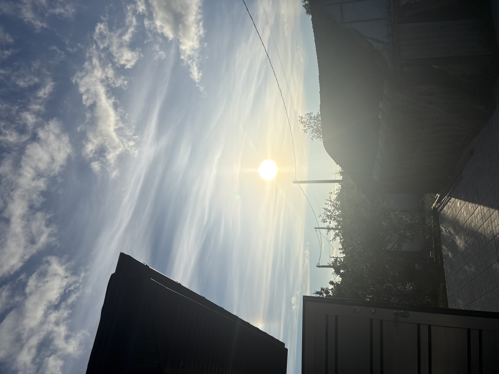 The image depicts an outdoor scene during daylight, likely in the city of Sovkhoza "Razdol'e", Russia. The perspective is from ground level, looking upwards. The sky is predominantly blue with wispy white clouds stretching horizontally across the frame. A bright, circular sun is visible in the upper center of the image, creating a lens flare effect and a slight green halo. In the foreground and to the left, the dark silhouette of a building's roof or overhang is prominent, with a section of a gray, horizontally-slatted gate or door visible on the far left. To the right, another dark-roofed structure, possibly a shed or garage, is partially obscured by sunlight. Trees and vegetation are visible in the mid-ground behind these structures, with power lines running across the sky. The ground in the foreground is paved with light-colored rectangular tiles, showing shadows cast by the buildings. The overall lighting suggests either early morning or late afternoon. There are no people or animals visible in the frame.