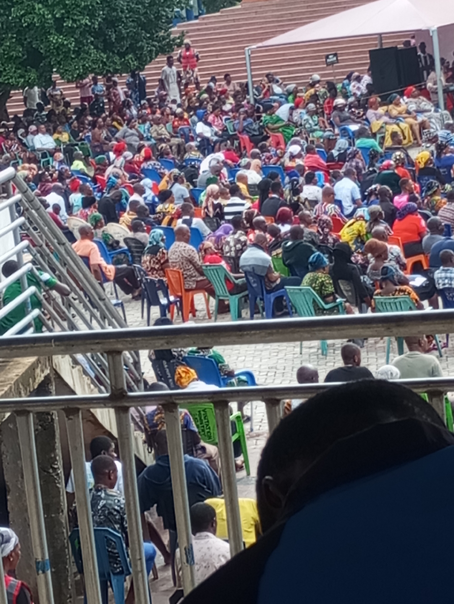 A large crowd of people is gathered in Onitsha, Nigeria. The people are seated in rows of plastic chairs in an outdoor setting. Above the crowd is a tiered seating area, also occupied by people. A metal railing separates the foreground from the main gathering. Some trees are visible in the upper left of the image. A white canopy structure is present in the background. The crowd is diverse in attire, with many individuals wearing colorful fabrics.