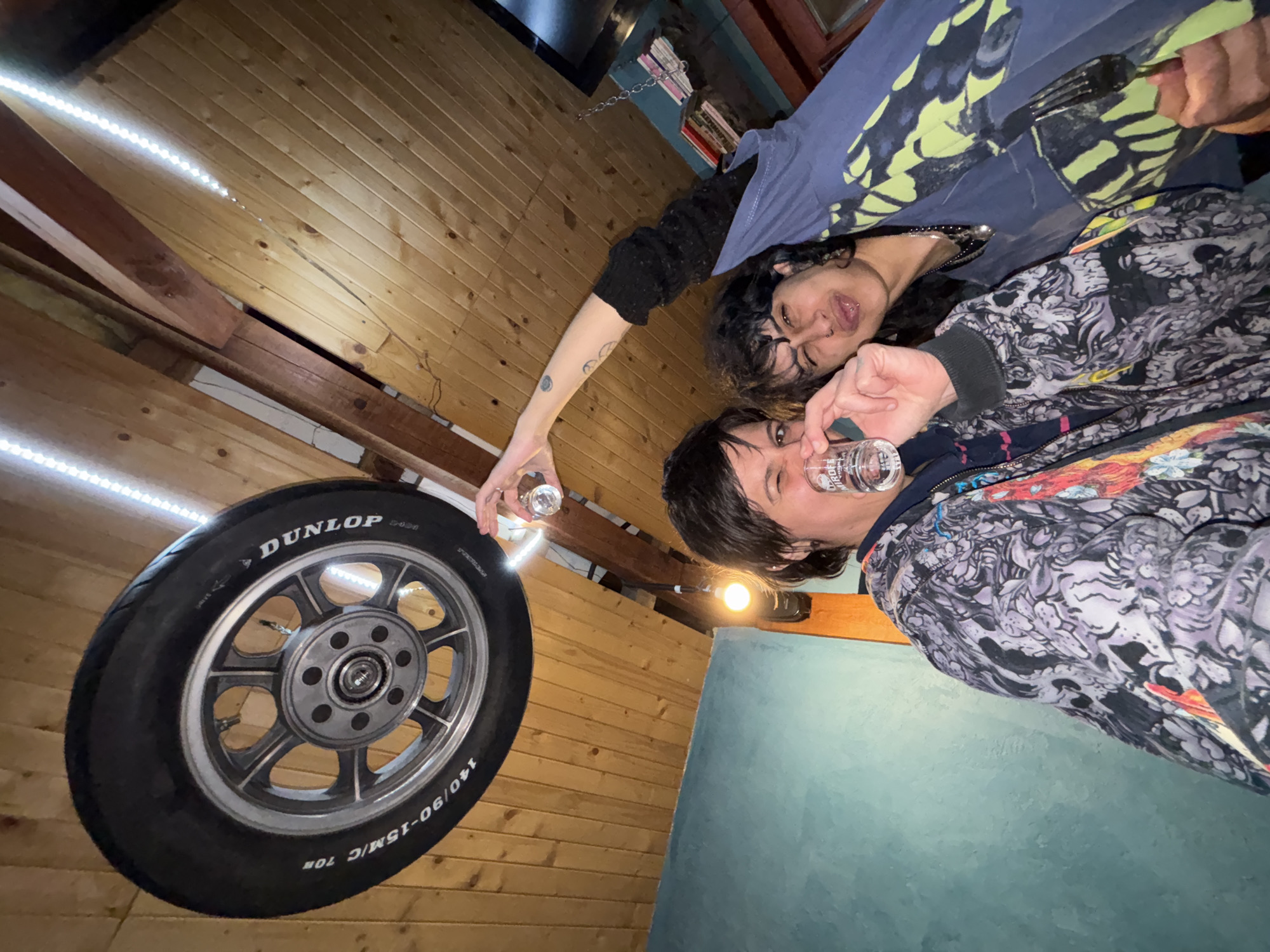 An overhead or low-angle, rotated shot captures two individuals in an indoor setting in Verinsko, Bulgaria. The background features wooden plank walls or ceiling, illuminated by a strip of LED lights running along a wooden beam. Another light fixture is visible, and in the upper right background, a shelf with books and a decorative chain can be seen. A light blue-green wall surface occupies the bottom right of the image.
On the right side of the frame (original orientation), one individual with dark, short hair is holding a shot glass to their lips, looking towards the camera with a casual expression. They are wearing a patterned hoodie, possibly with skull motifs. Closer to the left, another person with dark, curly hair, glasses, and a nose piercing has an arm extended towards a prominent tire, holding a second shot glass. This individual looks towards the camera with a playful, open-mouthed expression, with their tongue slightly visible. They wear a dark top and a blue and yellow patterned jacket.
The dominant object, aside from the people, is a large car or motorcycle tire with a wheel rim, prominently displaying the text "DUNLOP" and "140/90-15M/C 70H". The artificial lighting from the LED strips and bulbs suggests the photo was taken at night or in a dimly lit indoor space, possibly a rustic bar or a uniquely decorated room. The expressions and shot glasses imply a casual, social, and possibly festive atmosphere.
