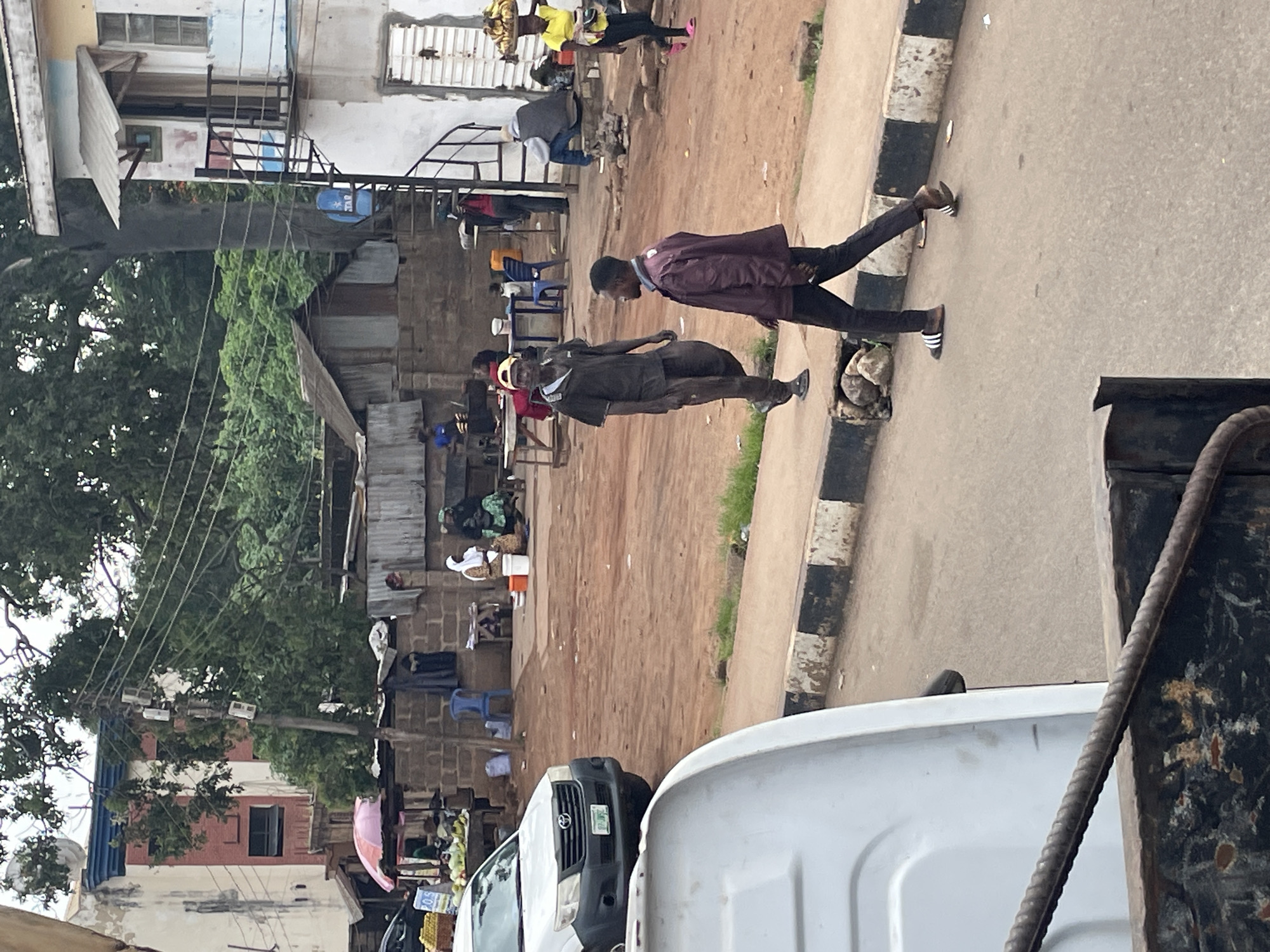 This is a street scene in Jos, Nigeria, with a focus on pedestrians and the urban environment. In the foreground, a white van is partially visible. Two men are walking on the sidewalk in the middle ground. The man on the left is wearing a dark t-shirt, dark pants, and a red cap, and appears to be looking to his right. The man on the right is wearing a maroon jacket and dark pants, and is walking away from the camera. Behind them, a white car is parked near a market stall with produce and a pink umbrella. Further back, several people are visible near buildings, some sitting and some standing. The environment appears to be a mix of informal market activity and residential or commercial structures, with overhead power lines and trees present. The ground is a mix of dirt and pavement.