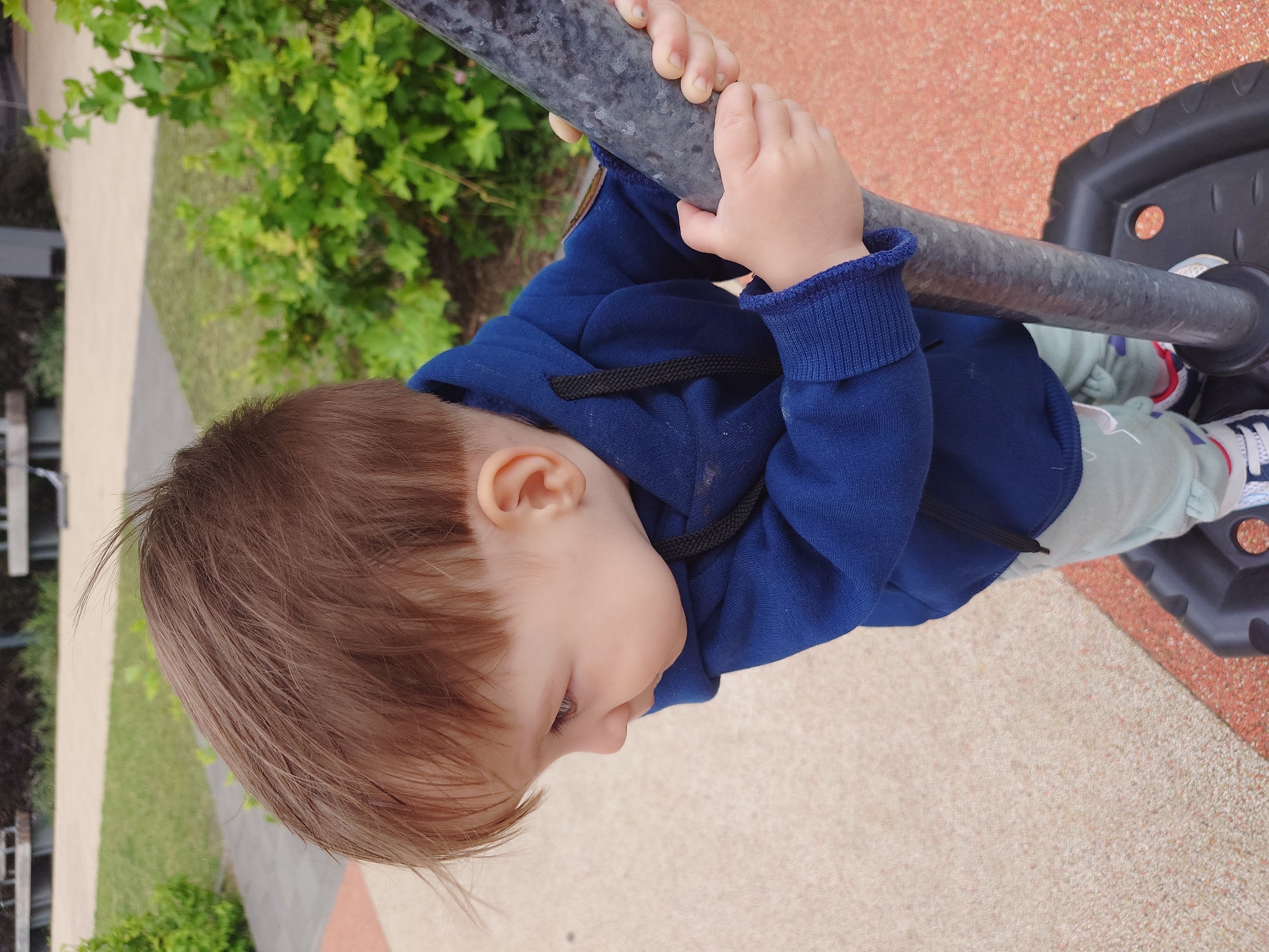 In an outdoor setting, a young child with brown hair is viewed from a high angle, looking to their left. The child is wearing a blue hooded sweatshirt and light-colored pants. They are holding onto a dark gray, textured pole with both hands, their fingers wrapped around it. The background consists of a grassy area with green bushes, a textured ground with orange and beige tones, and some blurred-out structures in the distance, possibly benches. The scene suggests a park or playground environment. The location is identified as Kommunarka, Russia.