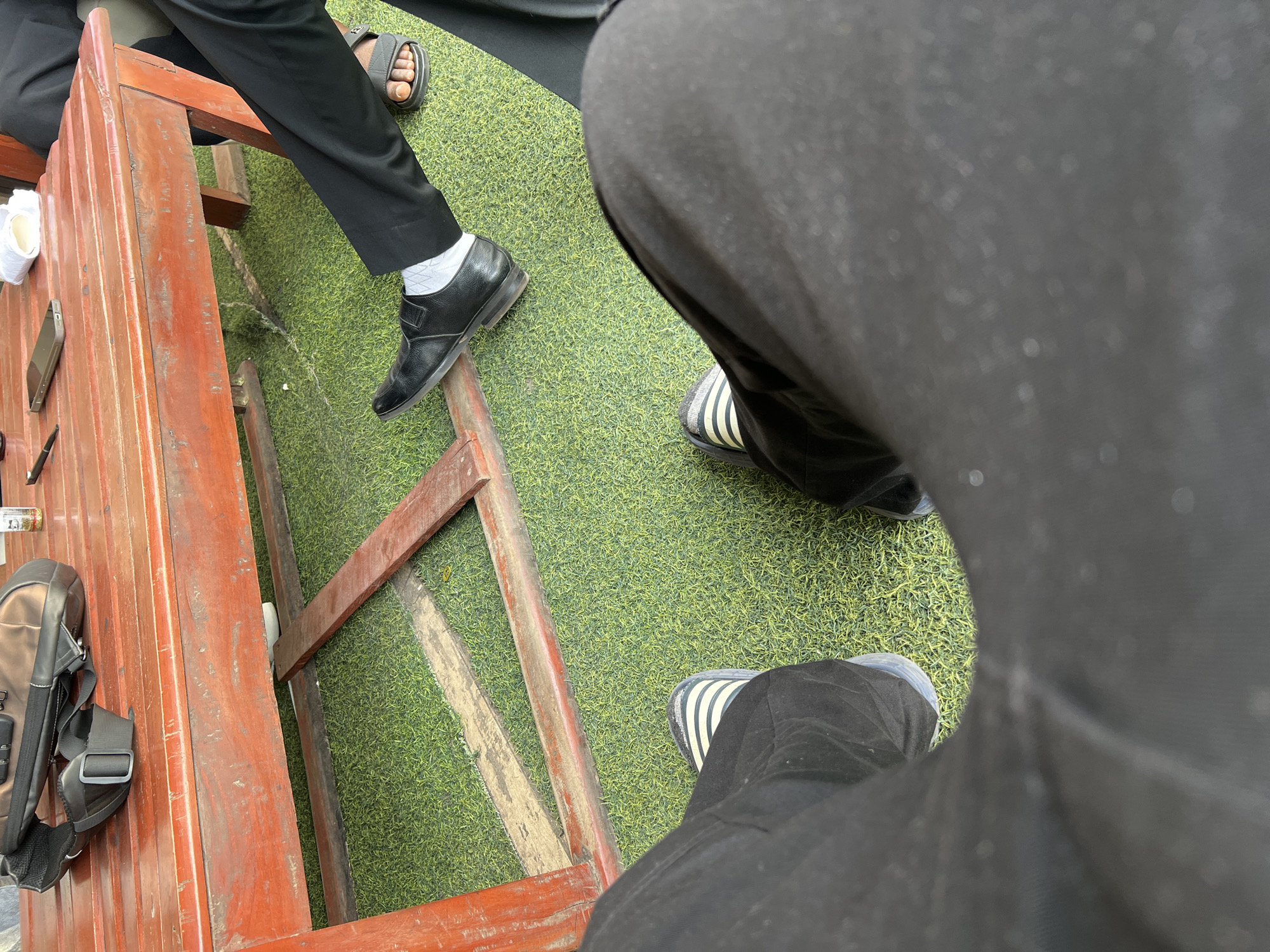 The image presents a top-down, slightly oblique view of a casual gathering of individuals, focused primarily on their legs, feet, and the immediate surroundings. The setting features a bright green artificial grass turf covering the ground, which also shows sections of a reddish-brown wooden frame or structure.
In the foreground, a person's legs are prominent, clad in black pants, with their feet visible in black and white striped slides. To the left, another individual is seated at a sturdy, reddish-brown wooden table or bench made of planks. This person is wearing black trousers, a black leather dress shoe with white socks on one foot, and a dark sandal on the other foot. Further to the right, a third person's feet are visible, also wearing black and white striped slides.
On the wooden table, several personal items are placed: a silver-grey smartphone, a black pen, a partially visible white cup or container, and a brown and black messenger-style bag with a strap.
The scene suggests a relaxed, daytime environment, likely an outdoor or semi-outdoor space such as a cafe patio, lounge area, or a garden setting in Abuja, Nigeria. The artificial turf and wooden furniture create an informal atmosphere for a casual meeting or social interaction. No specific activity beyond sitting and conversing can be definitively identified, and no text is visible in the image. The perspective is from one of the seated individuals looking down at the group and their belongings.