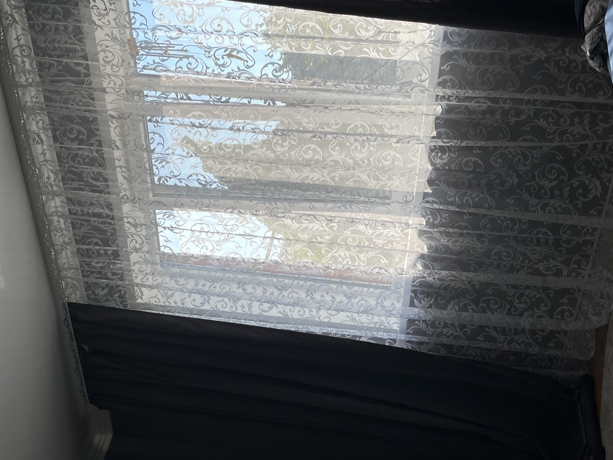 A medium shot shows a window covered by dark curtains on the left and a sheer, white, patterned curtain on the right. The window has multiple panes and is lit by sunlight. Outside the window, trees and a blue sky are visible. The overall impression is of an indoor scene looking outwards. The image does not contain enough visual information to determine its location in Saratov, Russia.