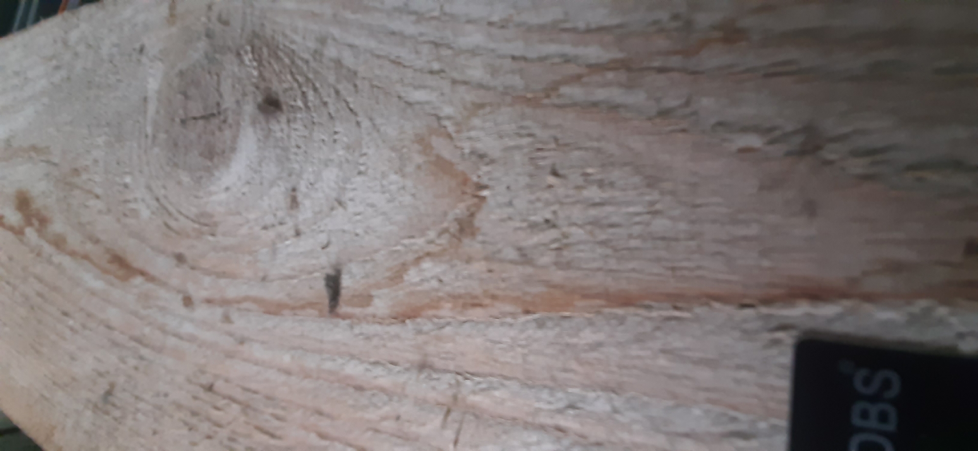 The image is a close-up shot of a light-colored wooden plank with visible grain patterns and a knot. The wood appears to be unfinished and textured. In the bottom left corner, a dark rectangular object with white lettering, partially obscured, is visible. The lettering appears to read "DBS". The background is the wooden plank itself, with variations in tone and texture due to the wood grain and knots. No people, animals, or specific landmarks within Yessentukskaya, Russia, are identifiable in this image. The focus is entirely on the surface of the wood and the partially visible object.
