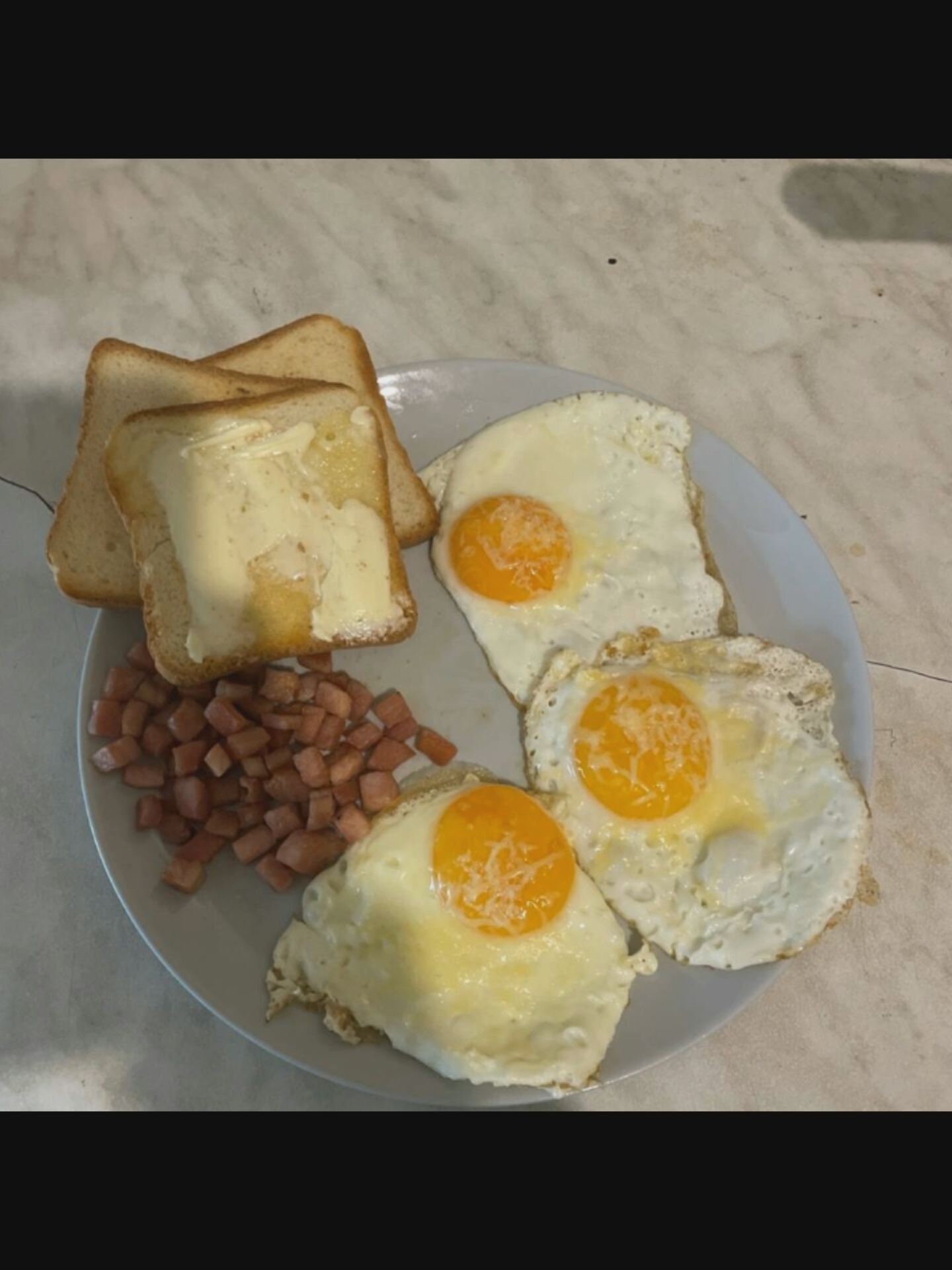 This media file shows a plate of food, likely a breakfast. The plate contains three fried eggs, two slices of buttered toast, and a pile of diced ham or bacon. The food is arranged on a light gray plate, which is placed on a speckled, light-colored countertop that appears to be marble or laminate.
The eggs are cooked sunny-side up, with visible yolks. The toast appears to be white bread, toasted and spread with butter. The diced meat is small and reddish-brown in color.
The lighting suggests it might be daytime, possibly morning, given the nature of the meal. There are no people visible in the frame, and the setting is a kitchen or dining area. No text is visible in the image. The overall presentation of the food is simple and unfussy. The image is cropped to focus on the food, with a dark area at the top and bottom suggesting it might be part of a larger video or photograph.