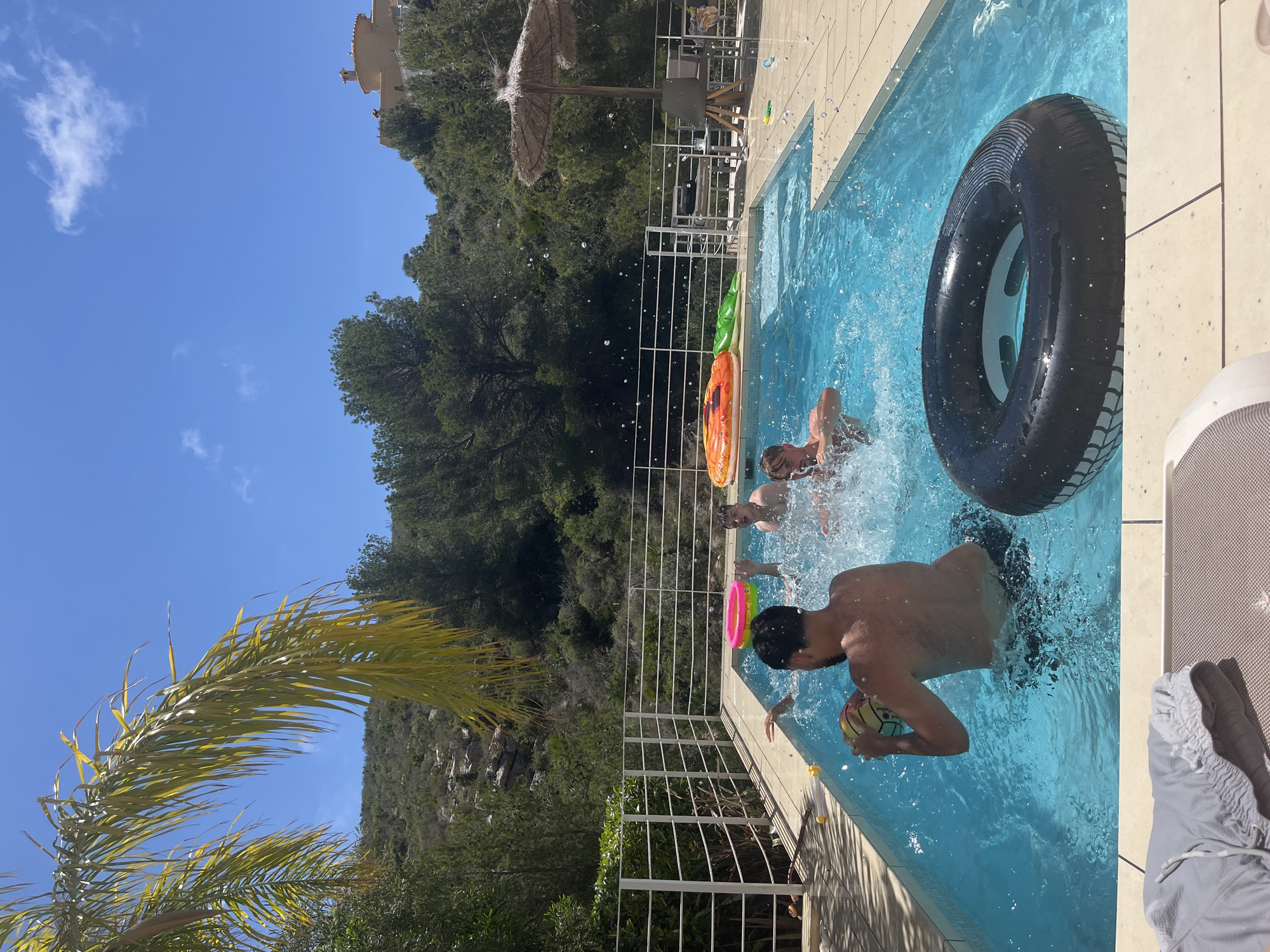 The image depicts a sunny day at a swimming pool, possibly located in a private residence in el Poble Nou de Benitatxell / Benitachell, Spain, based on the landscape visible in the background.
Three individuals are present in the pool, and a fourth person is partially visible in the lower right corner sitting next to the pool. One shirtless man holds a ball, and another person is visible near a black inner tube. Several inflatable pool toys are present.
The pool is surrounded by a tiled patio. A metal railing with a safety gate surrounds the pool. In the background, a hillside covered with trees and vegetation is visible, along with a building on top of the hillside. A sun umbrella is also visible next to the pool. A palm tree is visible on the left. The sky is clear with some clouds.