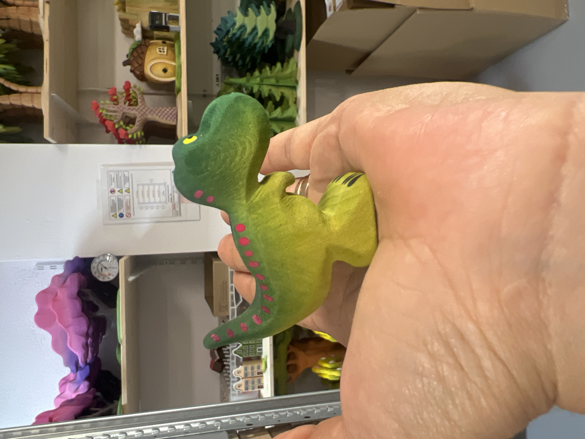 A person's hand holds a green and yellow toy dinosaur with pink polka dots. The dinosaur is positioned in the foreground, with its head facing right. In the background, shelves are visible, displaying various miniature items. These items include what appear to be colorful, abstract cloud-like sculptures on the top shelf, a miniature tree with red hearts on the second shelf, and a cluster of miniature evergreen trees on the third shelf. A cardboard box is also visible in the lower right portion of the image. The setting appears to be an indoor space, possibly a workshop or a retail display.