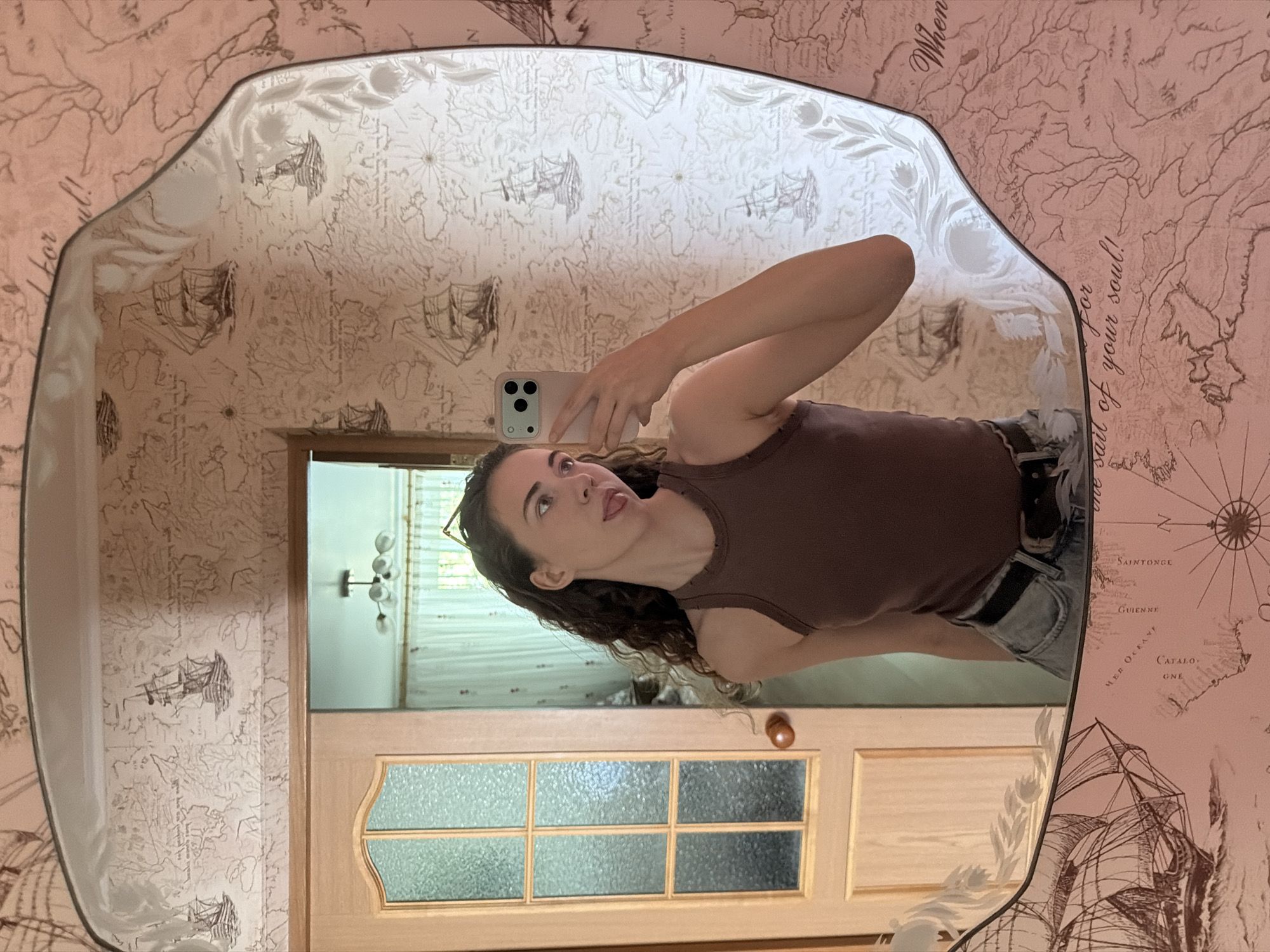 A young woman with long, wavy brown hair is taking a selfie in a mirror. She is wearing a brown tank top and blue jeans. She is sticking her tongue out at the camera. The mirror is framed with a pink wallpaper that has a map and ship design. The background shows a wooden door and a room with a chandelier. The location is the city of Zavodoukovsk, Russia.
