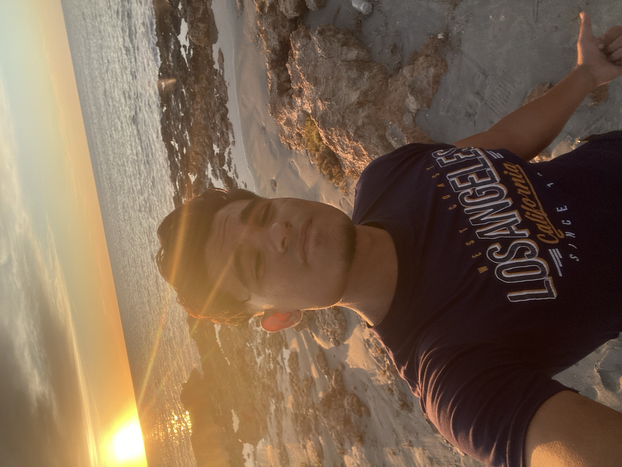 A medium shot angled selfie, taken at twilight, featuring a man on a beach. The man is facing the camera with his head cocked to the left, his right arm raised giving a thumbs up. He is wearing a dark blue t-shirt with “LOS ANGELES West Coast California Since” printed on the chest. The background shows a coastline with rocks, sand and water under sunset lighting. The scene appears to be located at a coastal area within the city of Juan Griego, Venezuela.