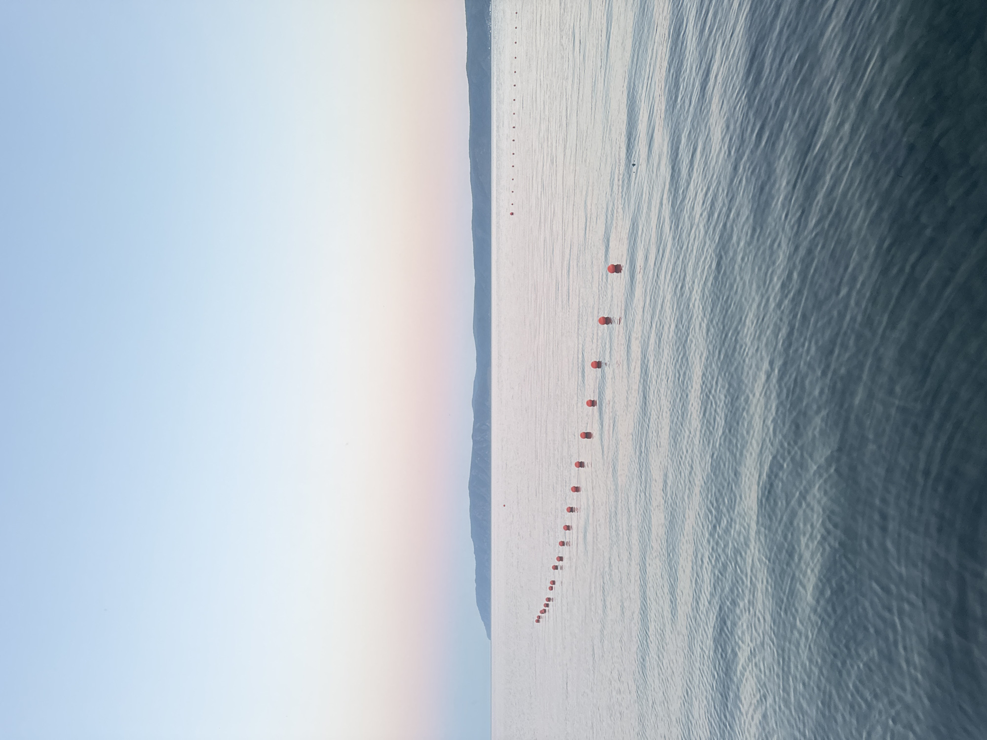 A serene seascape at dusk features a calm body of water with a gentle ripple pattern. In the foreground, a line of bright orange buoys forms a curved boundary across the water. The water transitions to a soft, muted blue as it stretches towards a distant, hazy landmass. The sky above is a gradient of pale blues and soft pinks, indicative of the twilight hour. The scene is devoid of any human presence or activity, creating a tranquil and isolated atmosphere. The location is not identifiable as Listvyanka, Russia.