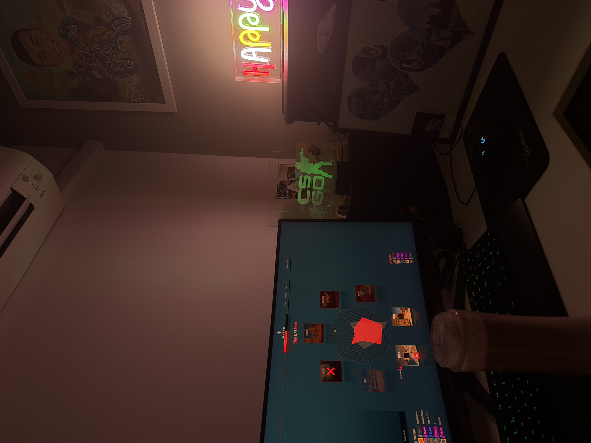 Here is a description of the image:
The image is a first-person view of a desk setup, primarily focusing on a computer screen displaying what appears to be a video game interface. The monitor shows a map selection screen with a variety of maps represented by small images. The text "Ban 0/1 Map" is visible, suggesting a map banning process in a game. Below the monitor, a portion of a keyboard is visible.
To the right of the monitor, there's an object that appears to be a cylindrical container. The background includes a poster of someone riding a motorcycle. There is a neon sign saying "Happy" in pink and yellow, and another light that is a green outline of Counter-Strike players and the initials CSGO. A mousepad and mouse are visible on a surface to the right of the monitor. The room is generally dimly lit.