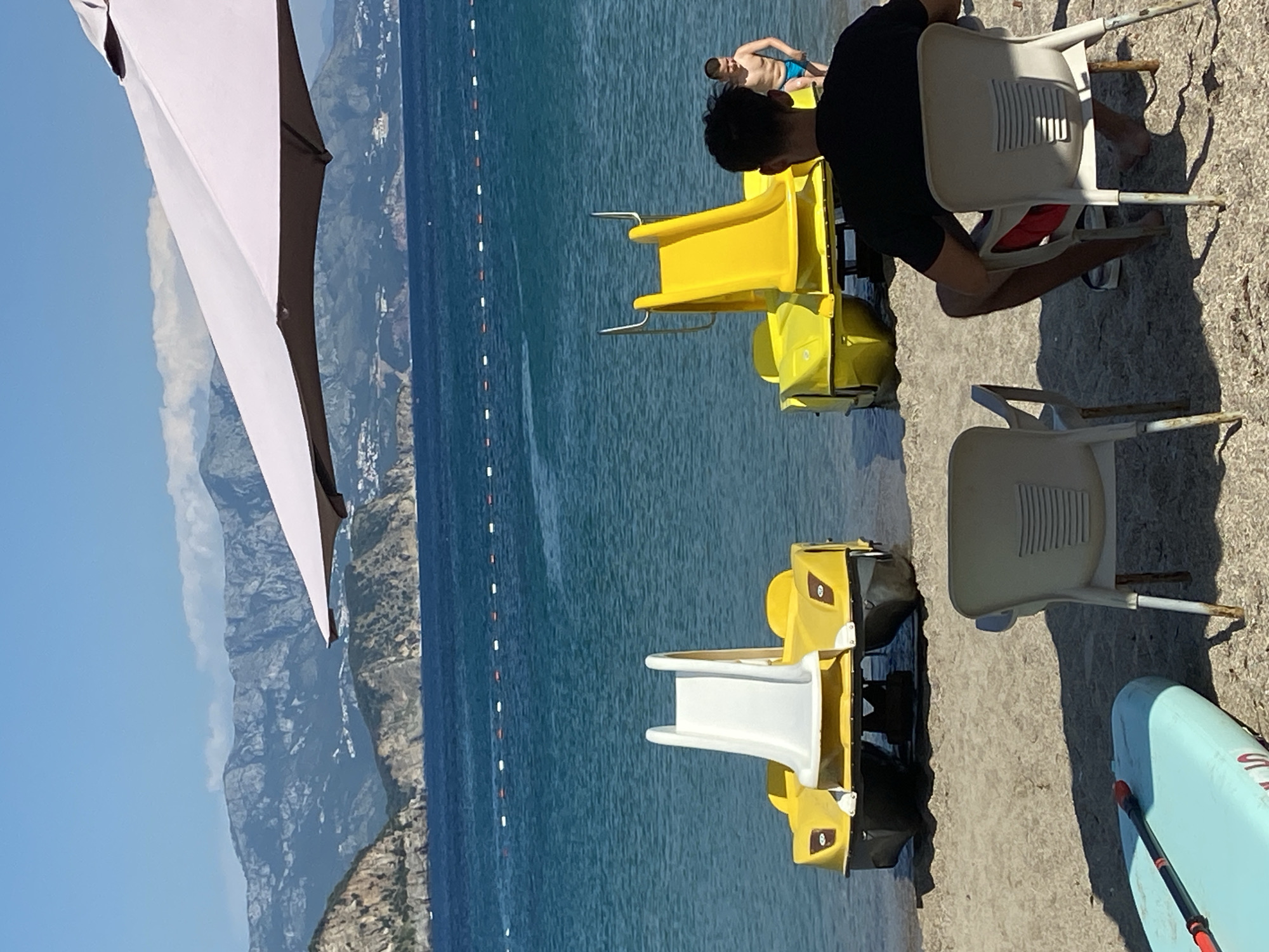 The image depicts a sunny beach scene in Prijevor, Montenegro, with the Adriatic Sea in the background. Two yellow pedal boats with slides are positioned on the sandy shore, facing the water. In the foreground, a man is seated in a white plastic chair, facing away from the camera, with his back to the sea. Another similar chair is positioned to his left. A light blue paddleboard with a red paddle lies on the sand in the lower left corner. A young boy in blue swim trunks stands near the right pedal boat, looking towards the water. Behind the boats, the clear blue sea stretches out, marked by a line of buoys. In the distance, a mountainous coastline with lush green vegetation and scattered buildings is visible under a clear blue sky. A large, light-colored umbrella shades a portion of the foreground.