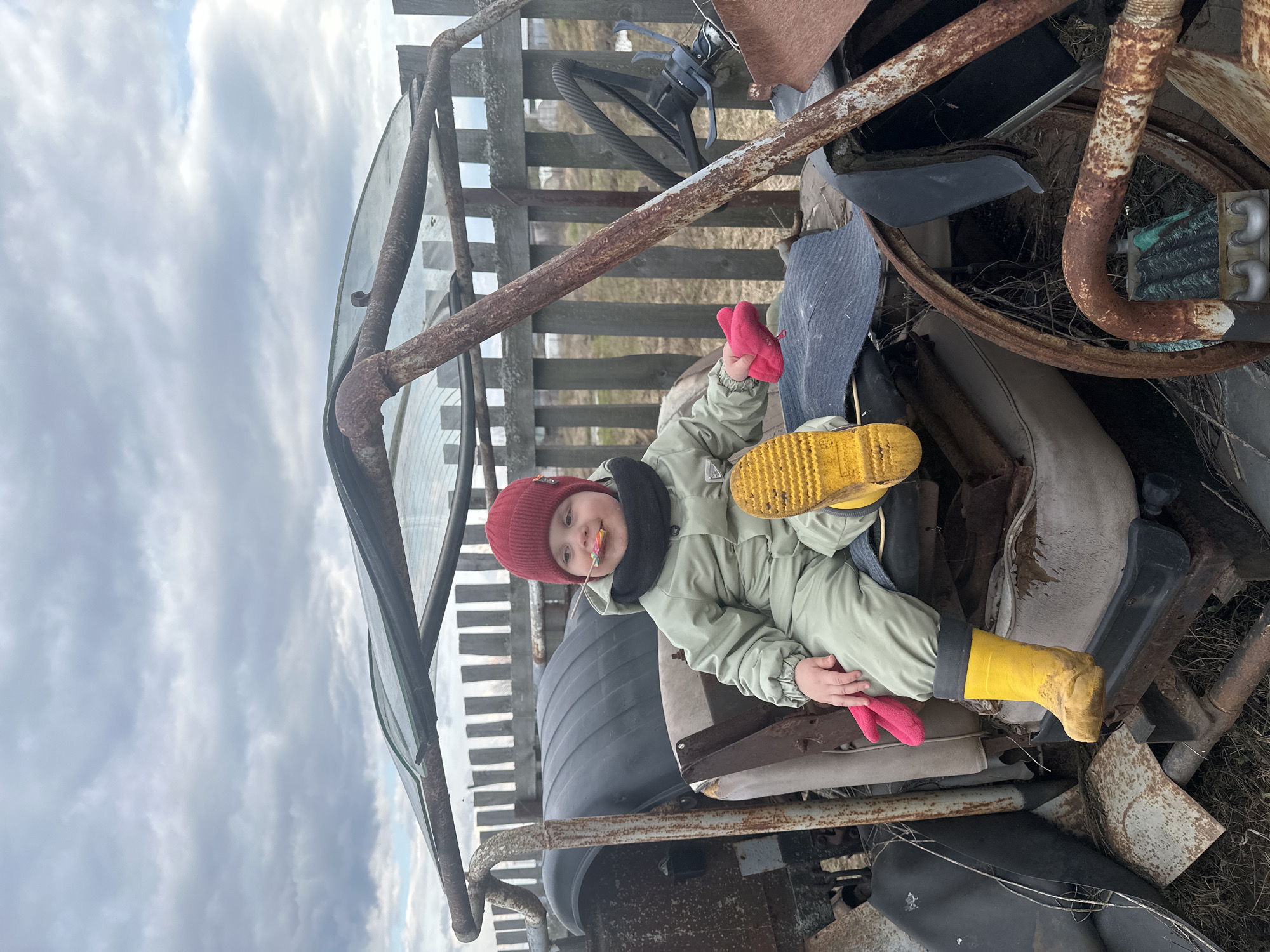 A young child wearing a red knit hat, a light green jumpsuit, yellow boots, and red mittens is sitting on top of a pile of discarded vehicle parts and other metallic debris. The child is holding a lollipop in their mouth. A rusty metal frame surrounds the child, and a wooden fence is visible in the background. The sky is overcast with grey clouds. No evidence indicates the location to be Ludorvay, Russia.
