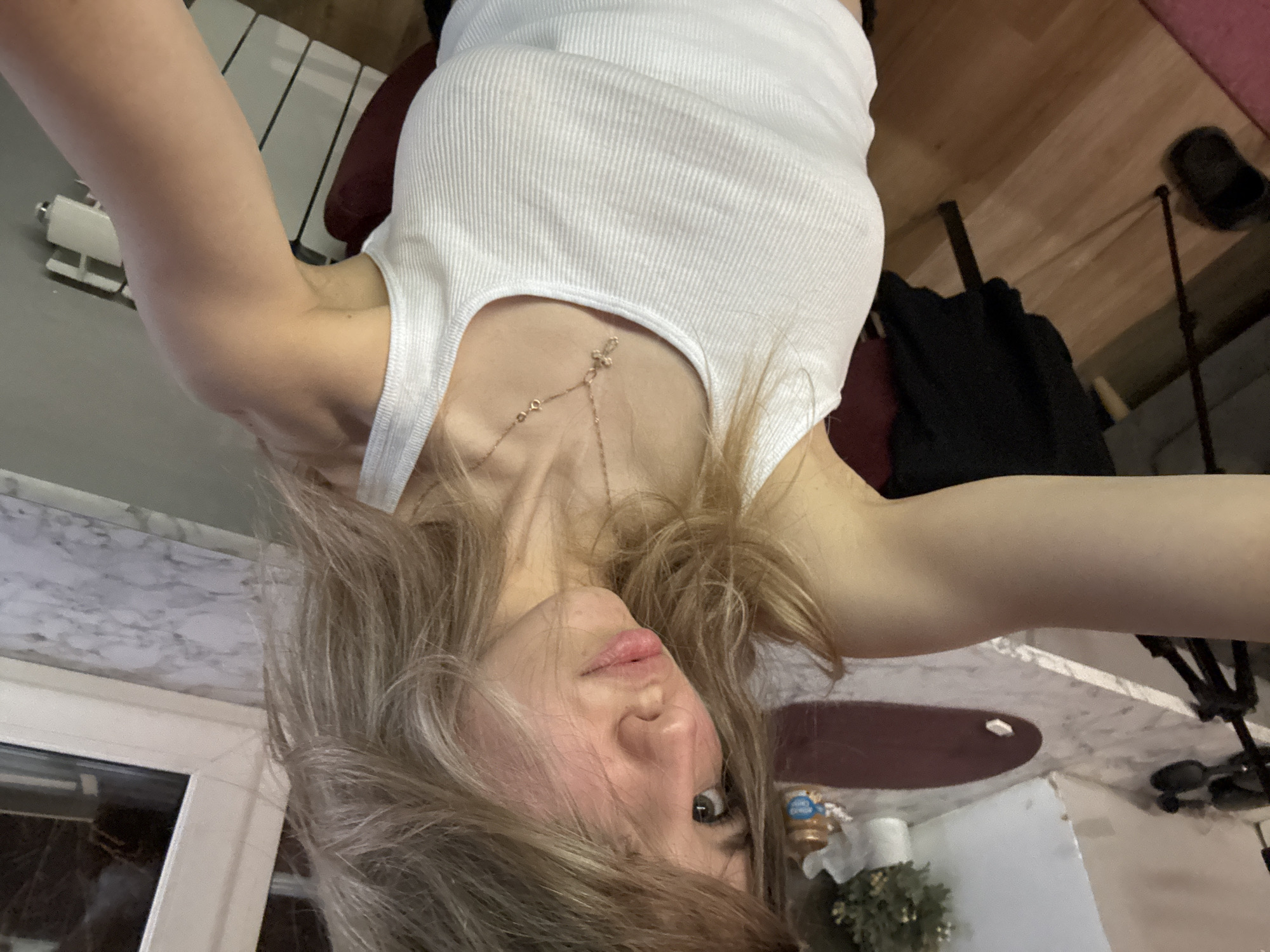 A young adult woman is seen in a self-portrait style photograph taken from an upside-down perspective, possibly with a mobile phone. The woman is wearing a white ribbed tank top and a delicate gold necklace. The background consists of interior details, including a marble-patterned surface, a partially visible window, a chair, and elements of a room setting. The location and city are not discernible from the image.