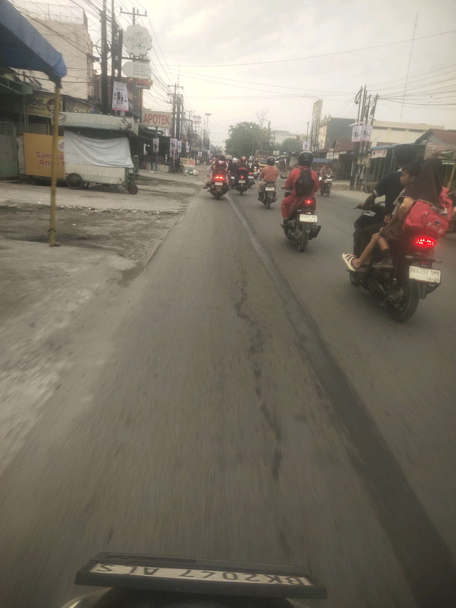 The image shows a street scene in Medan, Indonesia, with numerous motorbikes on the road. The perspective suggests the photo was taken from a moving motorbike. On the left side of the road, there are buildings with shops and signage, including "APOTEK" (pharmacy) and "ABOUT YOU." Several motorbikes are visible, some with riders wearing helmets and backpacks. One motorbike in the foreground has a red backpack and a license plate that reads "BK6392 AMV." The road is paved and appears to be a busy street. The sky is overcast. The overall impression is of a typical urban scene in Indonesia during daylight hours.