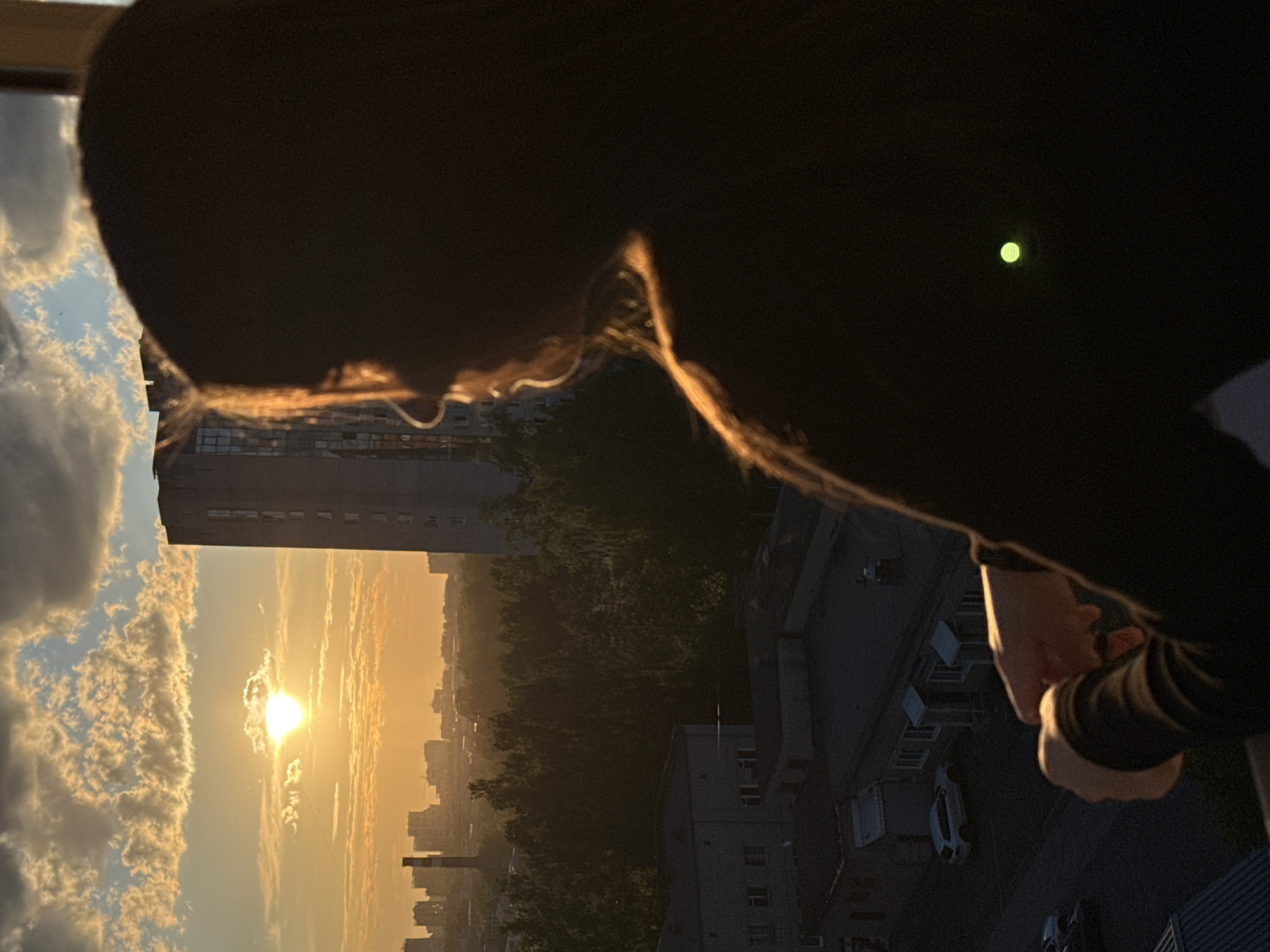 The image captures a scenic urban landscape in Izhevsk, Russia, viewed from a high vantage point during sunset. A person is prominently featured in the foreground, their back mostly silhouetted against the bright sky as they look out over the city.
The person's head, neck, and shoulder are dark, but strands of hair on their head and neck are illuminated with a golden glow by the setting sun, creating a distinctive outline. They appear to be wearing dark, long-sleeved attire, with a glimpse of their hands visible in the bottom right, also in shadow. A small, distinct green light is visible in the upper right portion of the dark foreground element, possibly an indicator light or reflection.
The sky is a central element of the scene, dominated by a brilliant sunset. The sun, appearing as a bright, slightly diffused orb, is low on the horizon. It casts warm orange, yellow, and golden hues across the clouds, which include scattered cumulus formations in the upper left and more elongated, streaky clouds further across the sky.
Below the horizon, the sprawling city of Izhevsk unfolds. Numerous buildings, mostly high-rises and other urban structures, are silhouetted or dimly lit against the receding light, stretching into the distance. A significant expanse of dark green trees, possibly a park or forested area, is visible between the buildings in the middle ground, adding depth to the urban fabric. The overall atmosphere is one of serene contemplation as day transitions to night over the bustling city. No visible text is present in the image.