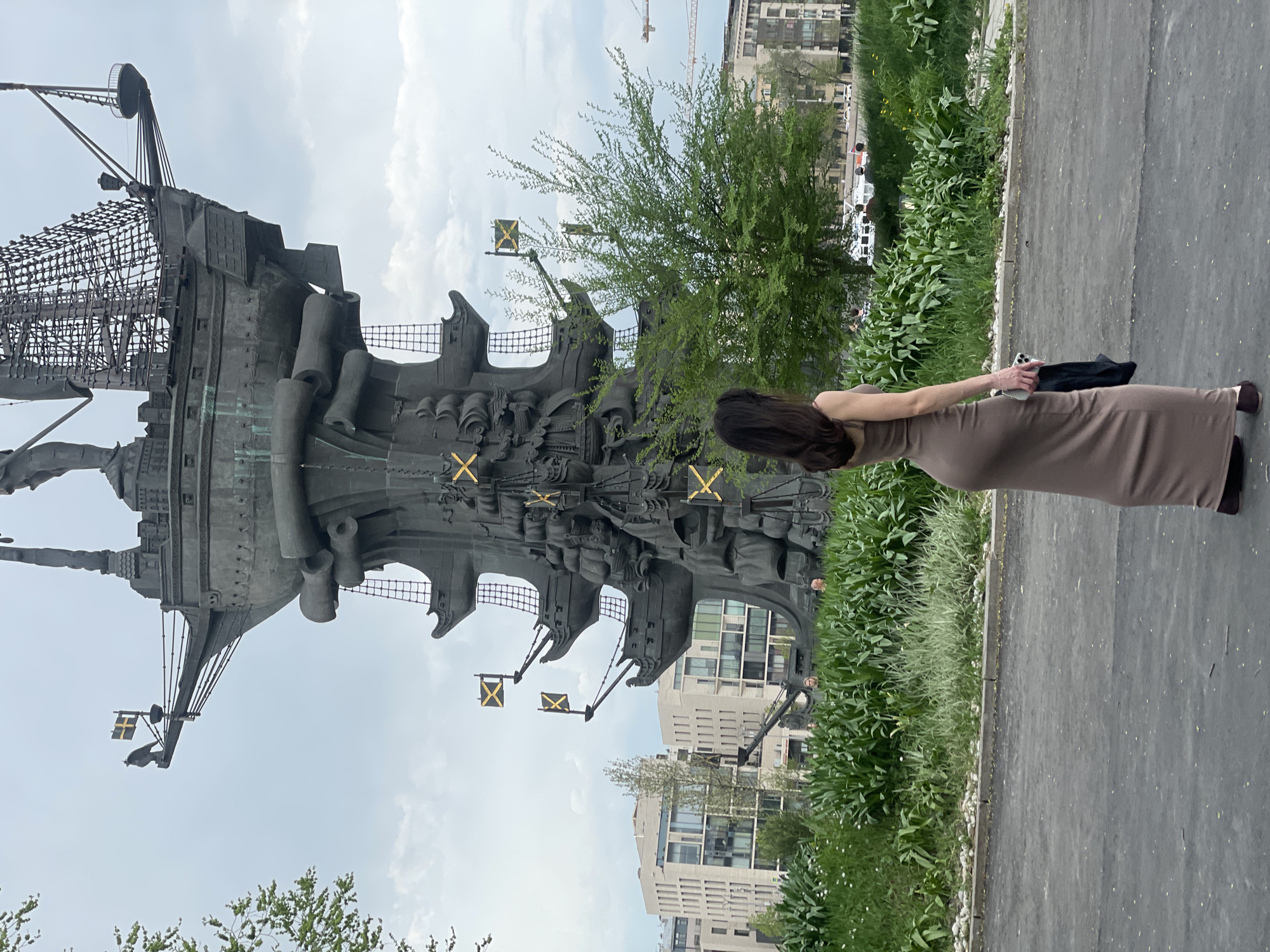 The image captures a bronze statue of a ship with masts and sails. A woman in a brown dress is standing to the right of the statue, facing it, and holding a phone in one hand and a black garment in the other. The statue has flags with crosses on it. Buildings are visible in the background. There is vegetation, including trees and grass, surrounding the statue and the woman. The scene appears to be outdoors on a paved area. The city is Unknown, Unknown.