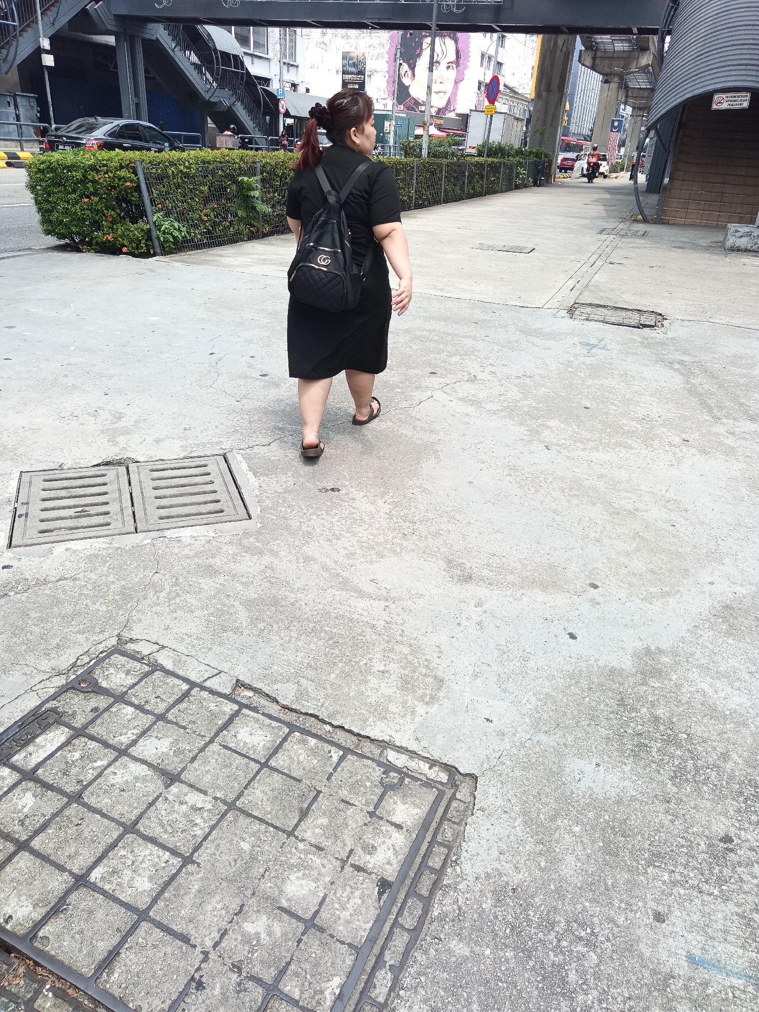 A woman in a black dress and a black backpack walks away from the camera on a concrete sidewalk in Kuala Lumpur, Malaysia. She is wearing brown sandals. In the background, there is a green hedge, a black car, an overhead walkway, and buildings. There are also two manhole covers on the sidewalk. The scene appears to be an urban environment with typical city infrastructure.