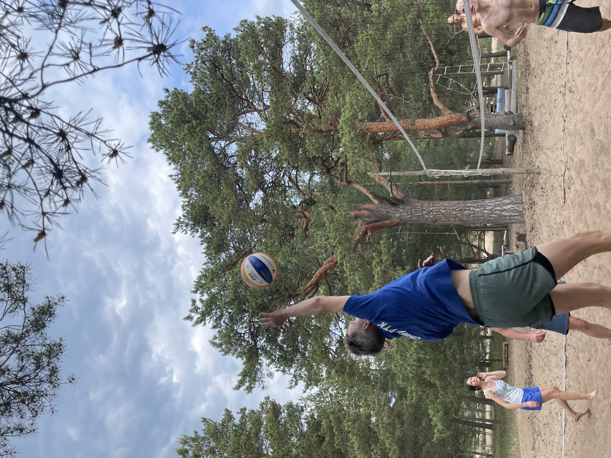 The image displays an outdoor beach volleyball game occurring on a sandy court within a pine forest in Zelenaya Roshcha, Russia, under a cloudy sky. In the foreground, a male player with short, light-colored hair is captured mid-action, wearing a blue t-shirt with Cyrillic text and dark green athletic shorts. He extends his right arm upward and forward, hand open, appearing to strike a blue, yellow, and white FIVA-branded volleyball suspended in the air slightly above and to his right.
To the left of the main player, a barefoot female with dark hair, wearing a white patterned tank top and blue shorts, stands on the sandy ground. She looks towards the volleyball play with her left hand positioned near her ear. The lower body of another player is partially visible between the main player and the woman.
A white volleyball net spans the middle ground, supported by two visible wooden poles. To the right of the net, a shirtless male wearing striped swimming shorts is partially visible, facing the net. Further into the background, a bald, shirtless male stands among the trees, observing the game.
The environment consists of light brown sand forming the court. Tall pine trees with dark green needles and reddish-brown trunks surround the court and extend into the background. Several pine branches with needles are visible in the extreme foreground, partially framing the upper portion of the scene. The sky is overcast, filled with light gray and white clouds. In the distant background, behind the net, wooden structures and a rope ladder are visible among the trees.