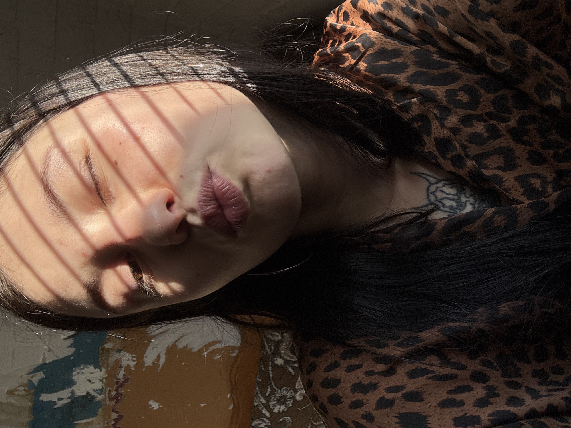 A medium shot photograph shows a woman's face bathed in harsh sunlight, obscured by the shadow of Venetian blinds. Her eyes are closed, and her lips are slightly pursed. She has dark hair that cascades around her face and onto a leopard print garment. Visible on her collarbone is a tattoo. The background is comprised of a partly visible floral rug and a peeling paint surface, suggesting an interior setting. The image is rotated counterclockwise by 90 degrees. There is no indication of the city Фоки, Russia in the image.