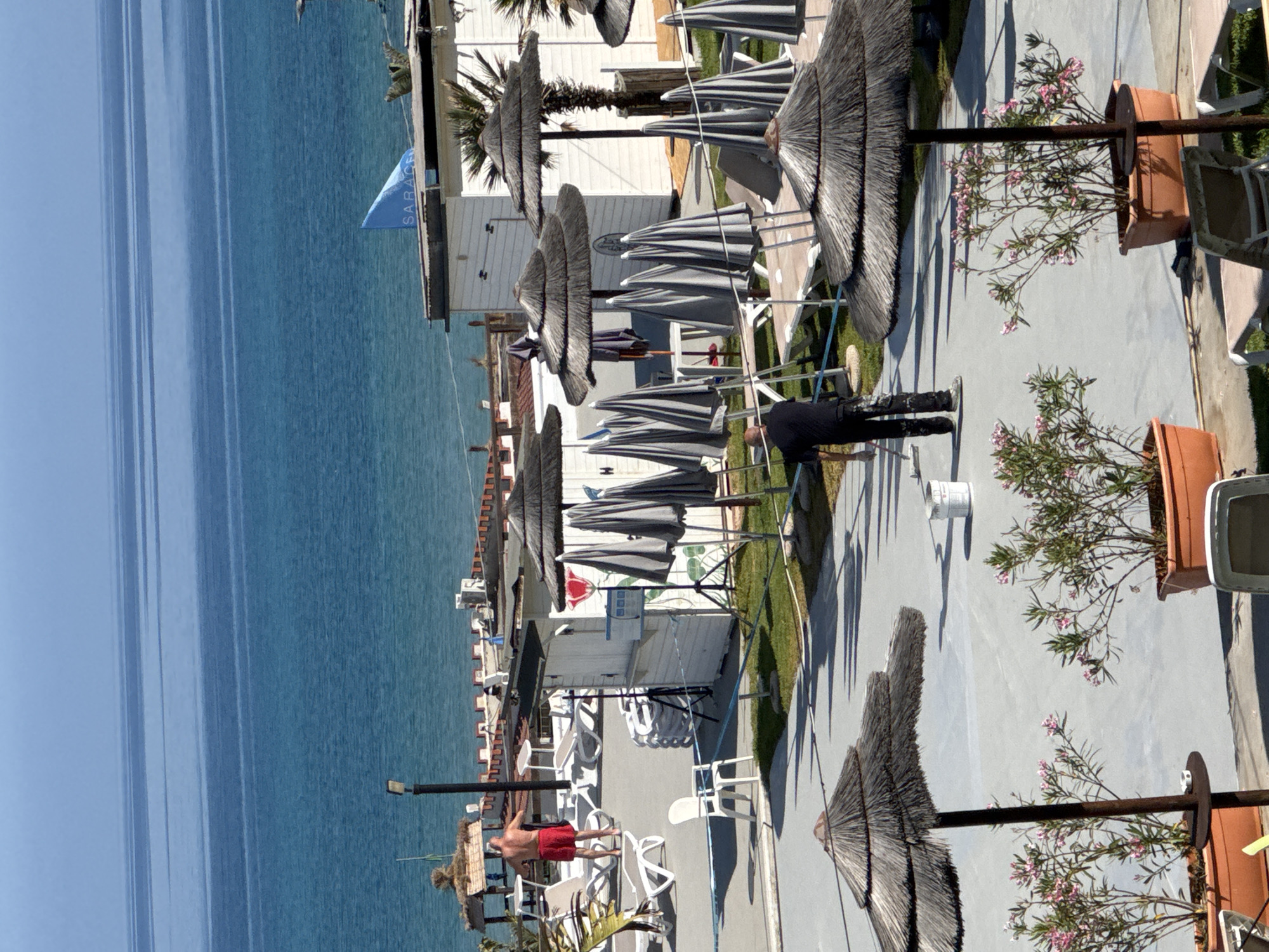 This image captures an outdoor scene at Lido Saracena in Isola delle Femmine, Palermo, Italy, under a clear blue sky. The Mediterranean Sea occupies the upper half of the frame, displaying various shades of blue. In the mid-ground, several white buildings and structures are visible, including a prominent white cabin with a blue flag bearing "SARACE" (likely part of "SARACENA") on its roof. A decorative low wall with crenellated tops separates this area from the sea.
On the left, a shirtless man in red swim trunks walks near several white sun loungers and a rustic hut with a thatched roof. A dark light pole stands nearby. In the foreground, a man dressed in black trousers and a dark shirt applies a light gray coating to a paved surface using a long-handled roller brush; a white bucket is situated near his feet. Numerous thatched beach umbrellas are scattered across the scene, some open and others folded. A cluster of gray, folded fabric umbrellas with dark poles stands upright near the white buildings. The paved surface is interspersed with patches of green grass and several terracotta pots containing green plants with small pink flowers, likely oleanders. A thin blue line, possibly a marker or rope, runs across the paved area. Long, distinct shadows indicate strong sunlight.