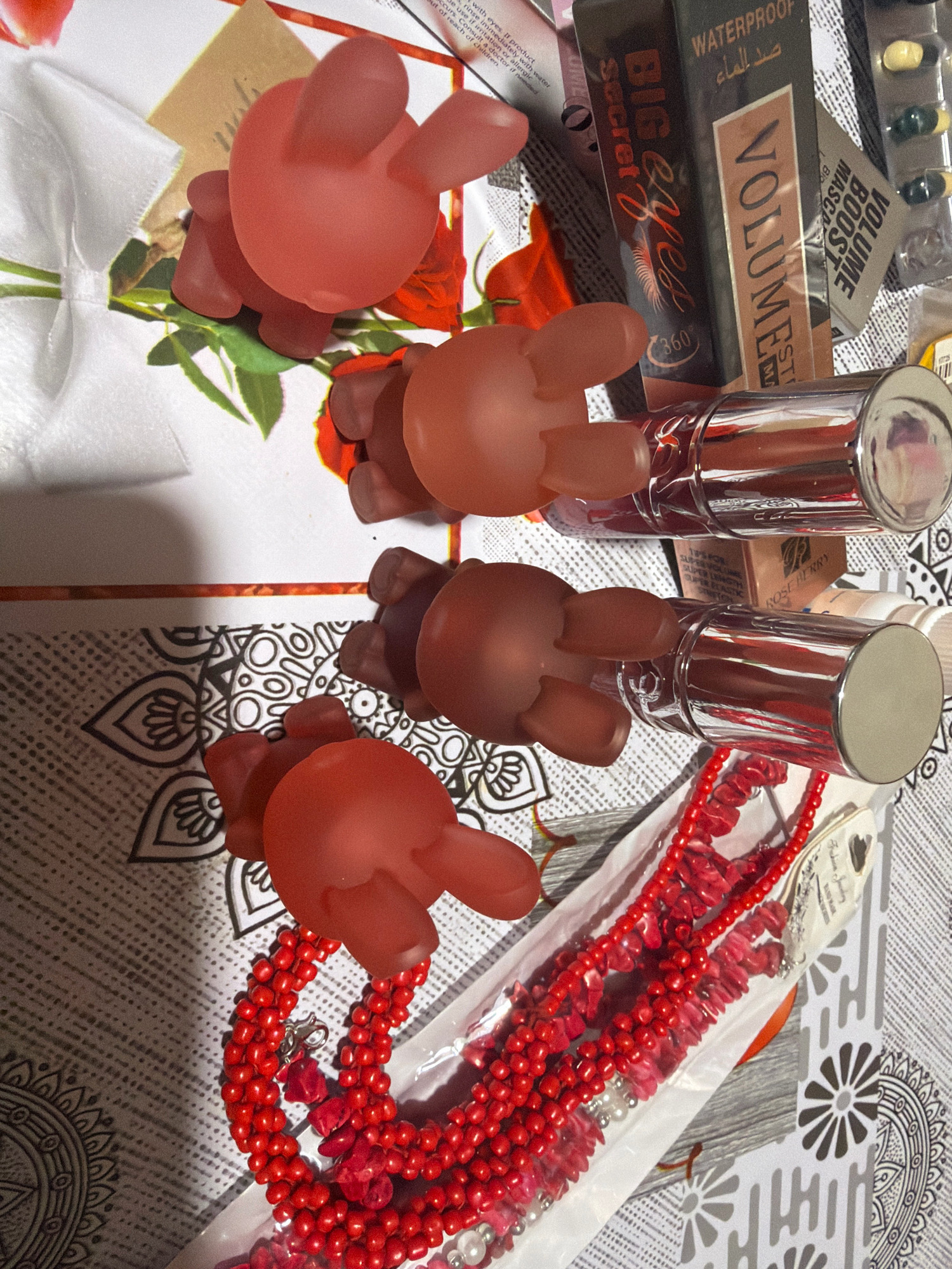 The image shows a flat lay of various items, including makeup products, decorative elements, and what appear to be toy figures. In the upper left, a white ribbon is visible. To its right, a red rose design is printed on a light brown background. Several pinkish-red, opaque toy figures resembling rabbits are positioned across the frame. Two lipsticks with silver caps are placed horizontally in the middle of the image. Below the lipsticks, a red beaded necklace is laid out, partially enclosed in plastic packaging. In the background, there are various patterns, including a black and white mandala-like design and a textured grey surface. To the far right, a small package containing pills or capsules is visible. The scene does not provide any information about the Municipal Unit of Nafplio, Greece, or any specific geographical location. There are no people present in the image.