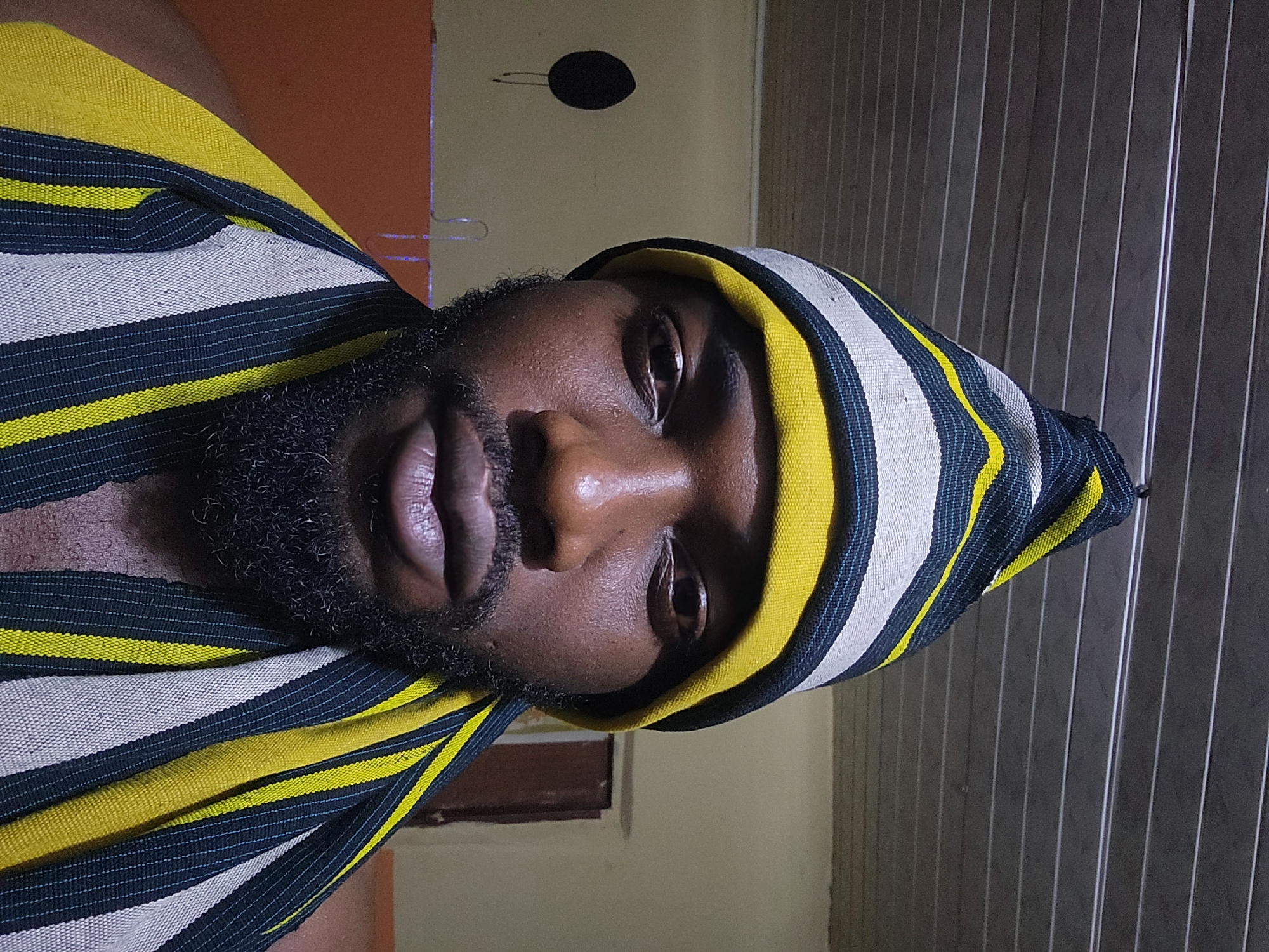A medium shot shows a Black man from the chest up, facing the camera. He is wearing a striped knit cap with yellow, white, and black stripes, and a matching striped scarf draped around his shoulders. The man has a beard and appears to be indoors, with a slatted ceiling visible above him. The background is a plain wall with some indistinct objects. The lighting suggests it is nighttime, and the overall environment appears to be a residential setting in Jos, Nigeria.