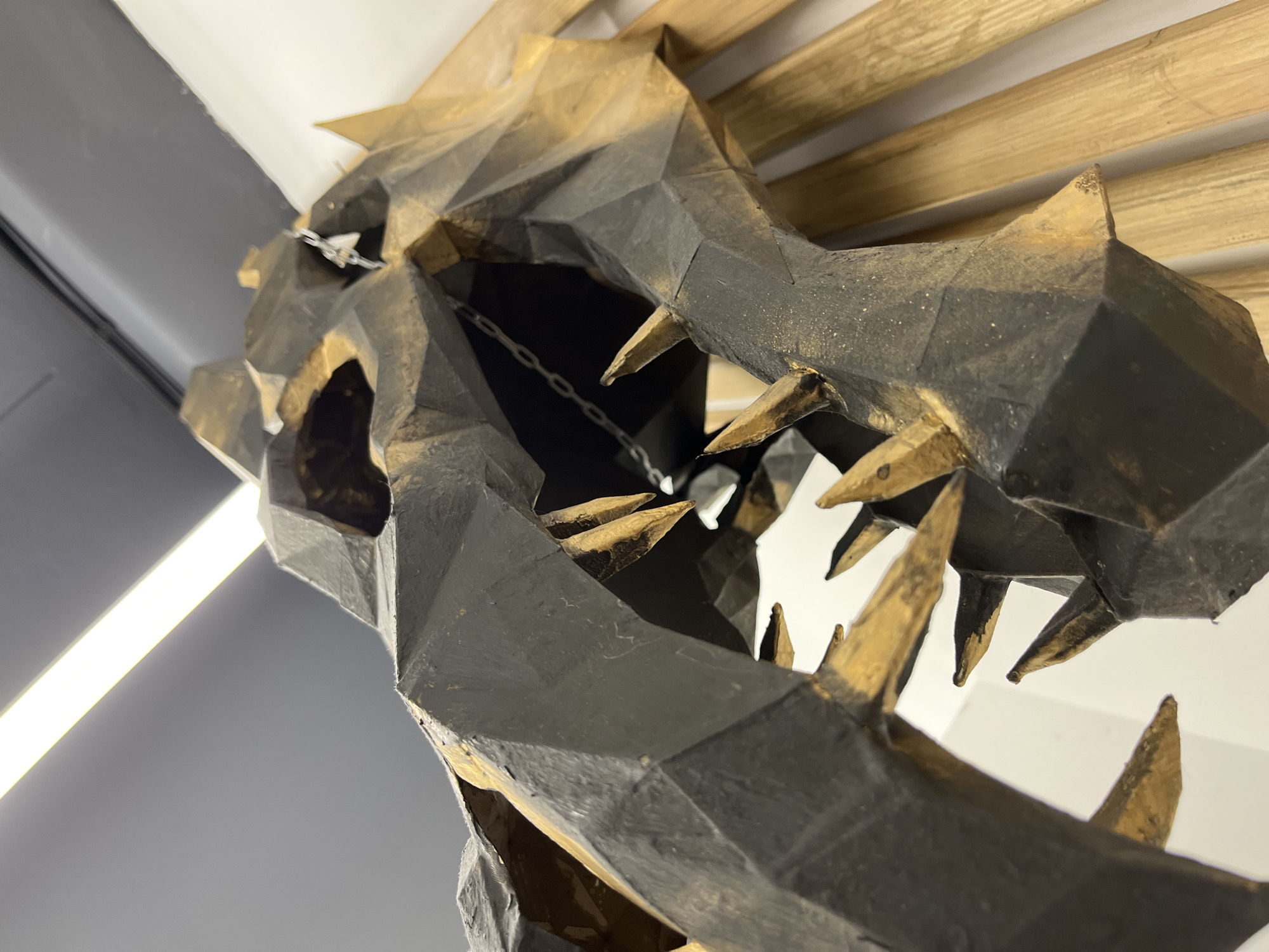 The image shows a low-poly model of a dragon's head, hanging from the ceiling. The head is constructed from geometric shapes, painted black with gold highlights. The mouth is open, revealing pointed teeth and a chain hanging inside. The background includes a gray wall, a fluorescent light fixture, and a wooden slat structure on the ceiling. The location is Chaikovsky, Russia.